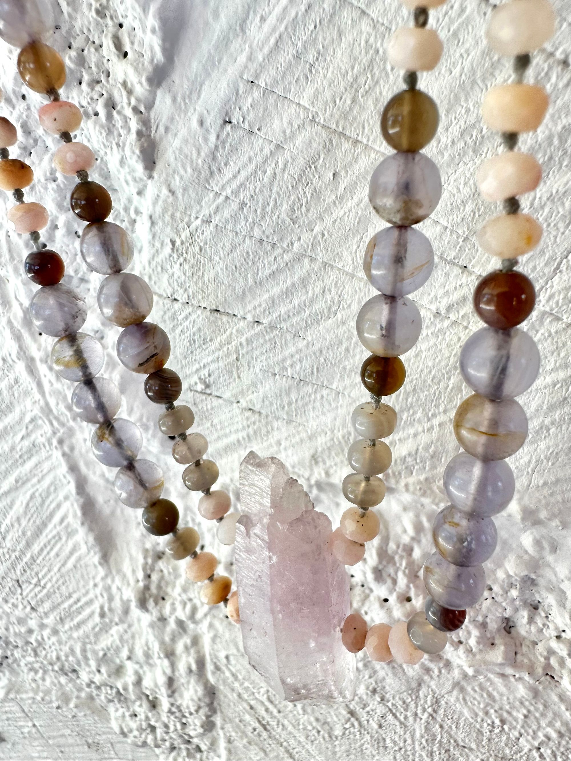 A necklace of soft but luminous, pink, purple and blue crystals hangs on a wall made of white painted cordwood