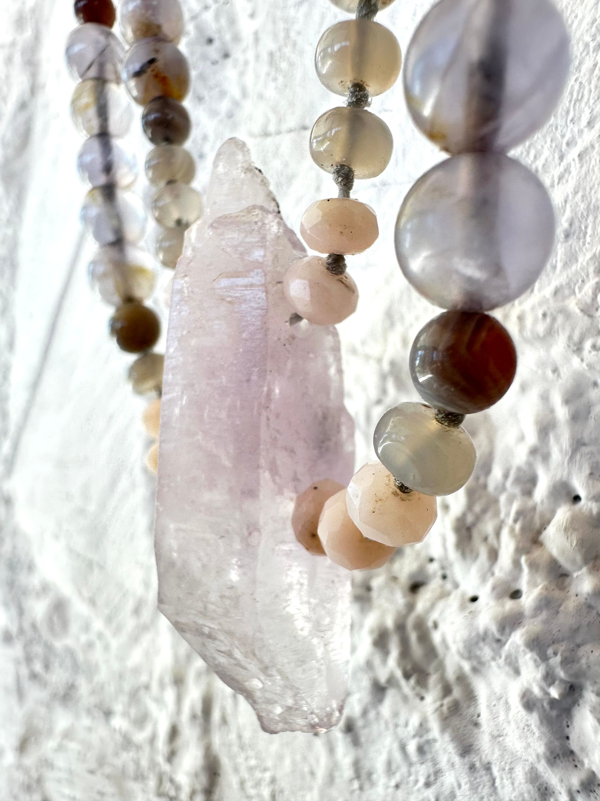 A necklace of soft but luminous, pink, purple and blue crystals hangs on a wall made of white painted cordwood