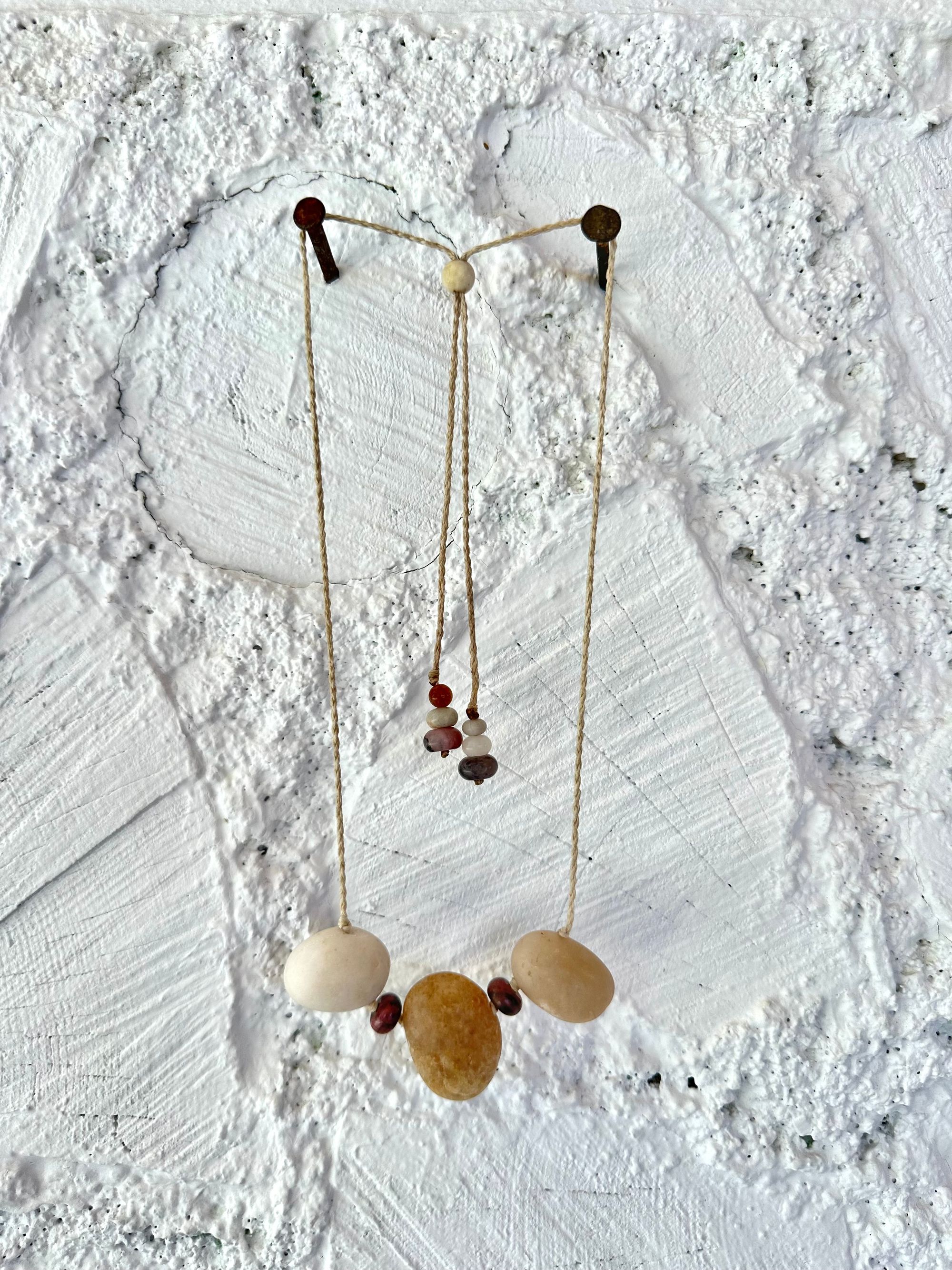 A simple necklace of three ocean tumbled stones that are tan and white with two pink beads of Jasper hangs from a white braided cord against a wall of white painted cordwood