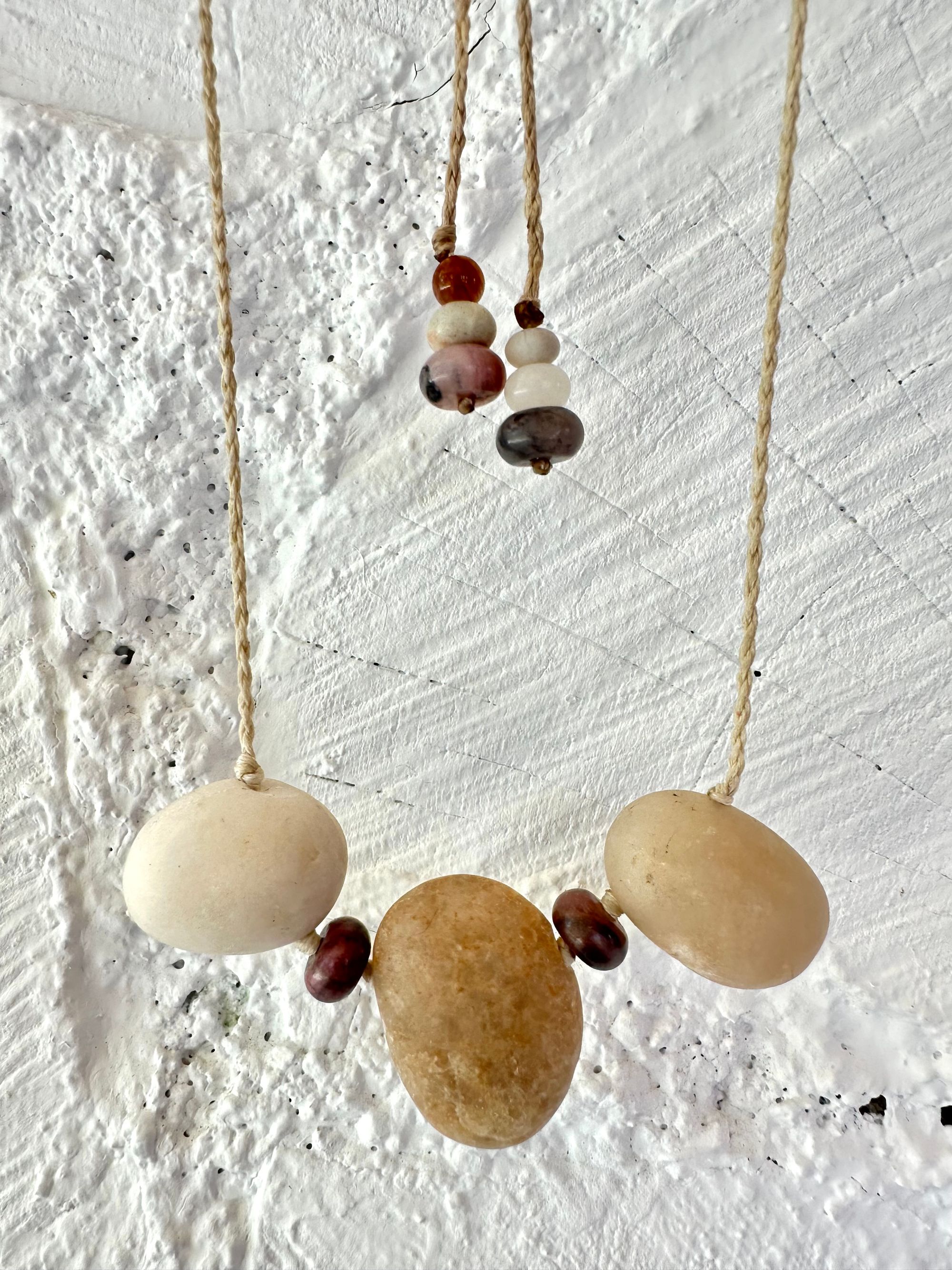 A simple necklace of three ocean tumbled stones that are tan and white with two pink beads of Jasper hangs from a white braided cord against a wall of white painted cordwood