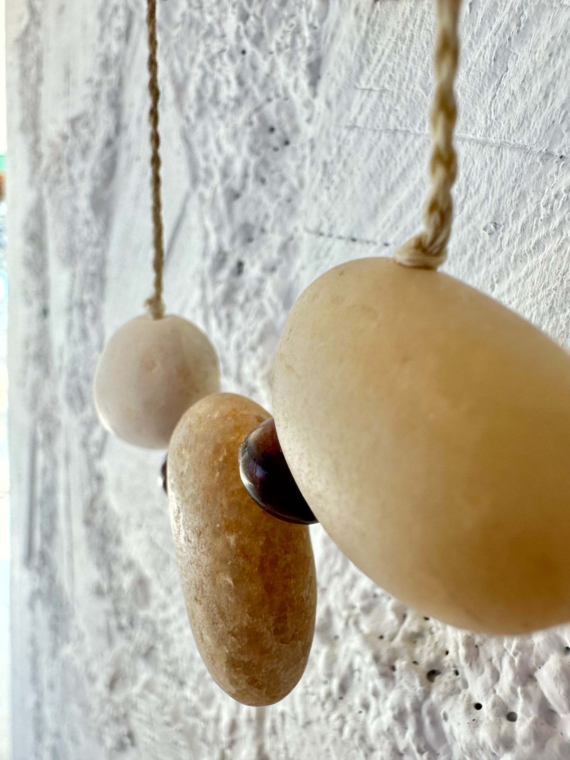 A simple necklace of three ocean tumbled stones that are tan and white with two pink beads of Jasper hangs from a white braided cord against a wall of white painted cordwood