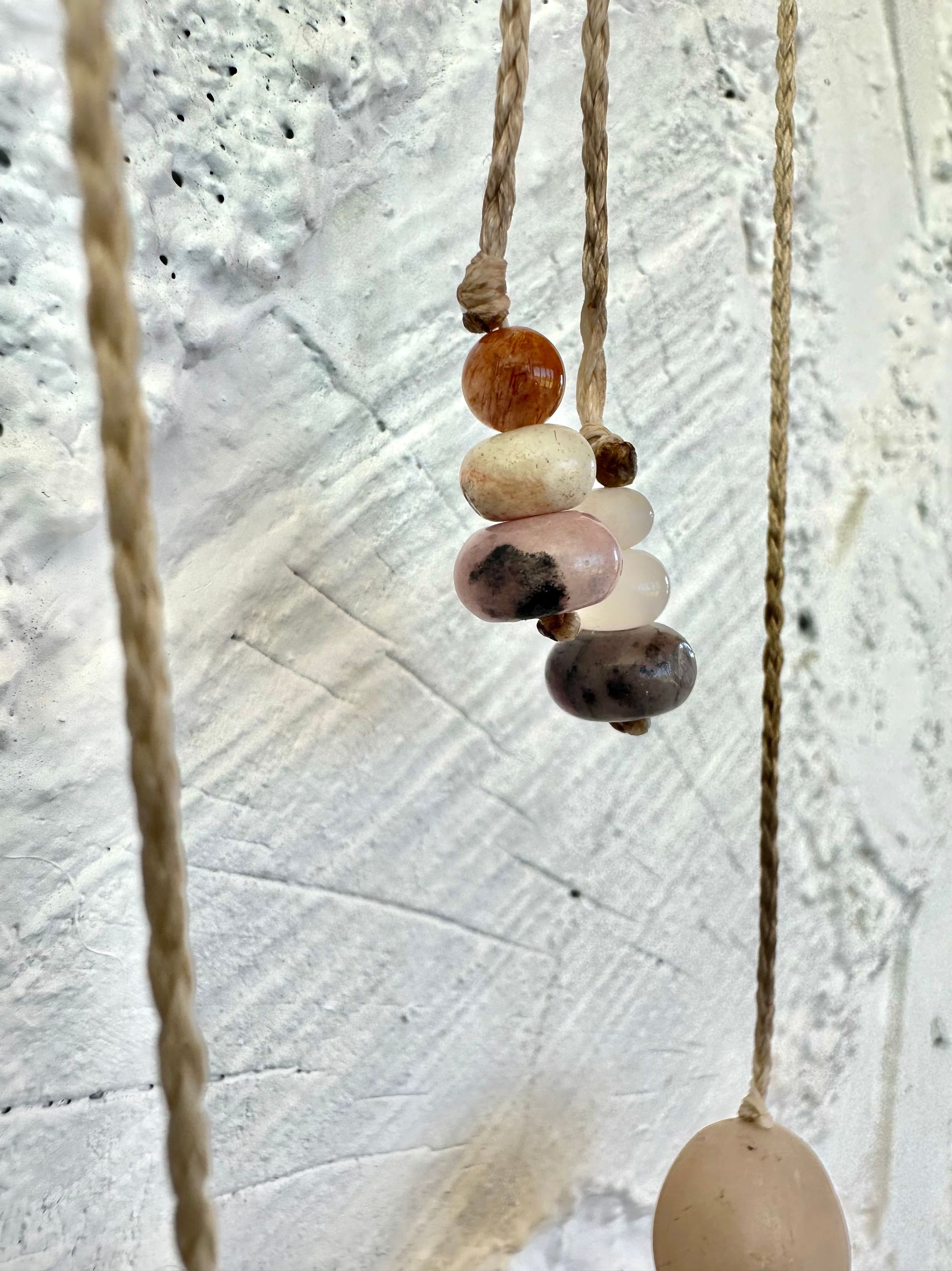 A simple necklace of three ocean tumbled stones that are tan and white with two pink beads of Jasper hangs from a white braided cord against a wall of white painted cordwood