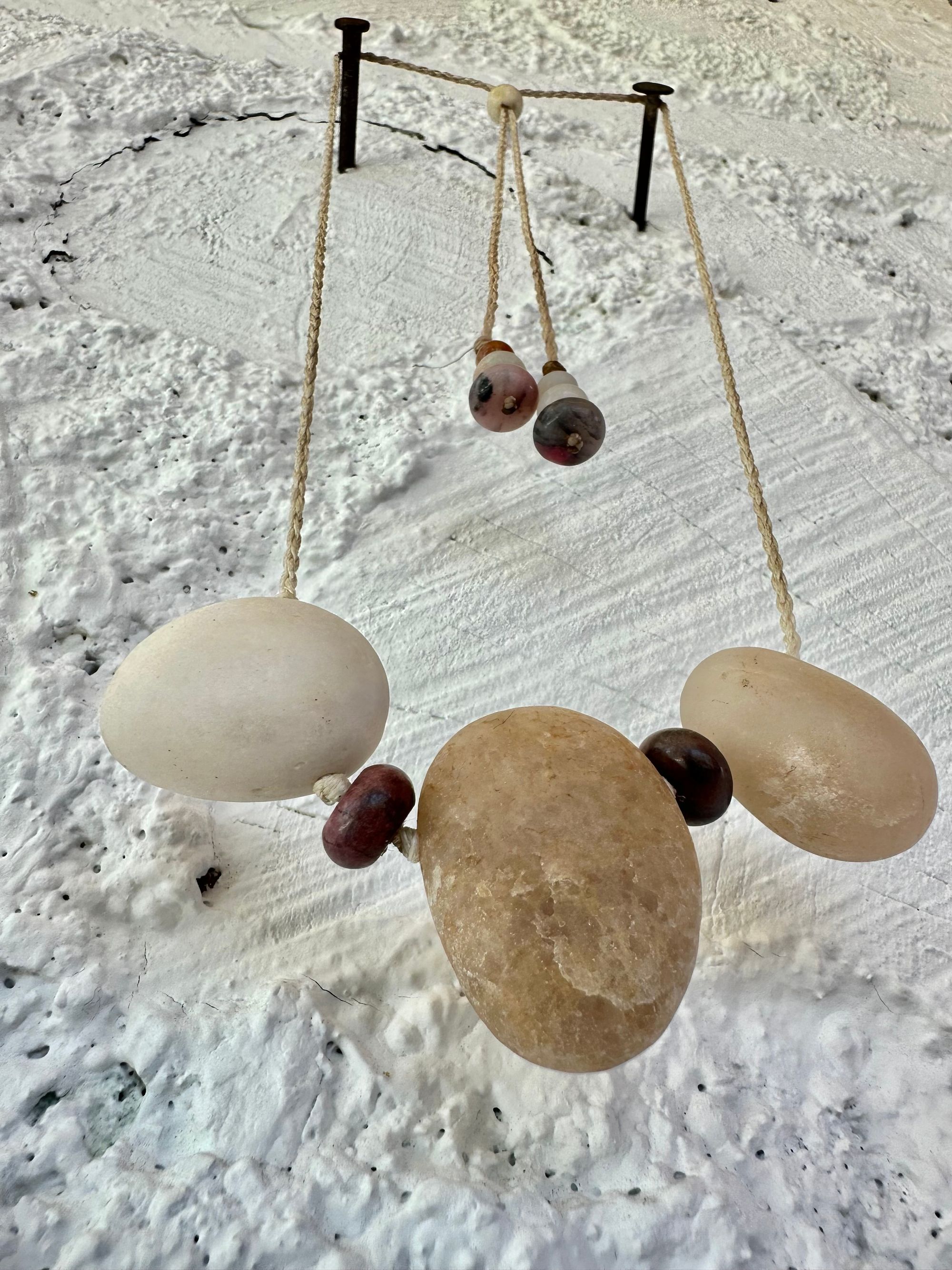 A simple necklace of three ocean tumbled stones that are tan and white with two pink beads of Jasper hangs from a white braided cord against a wall of white painted cordwood