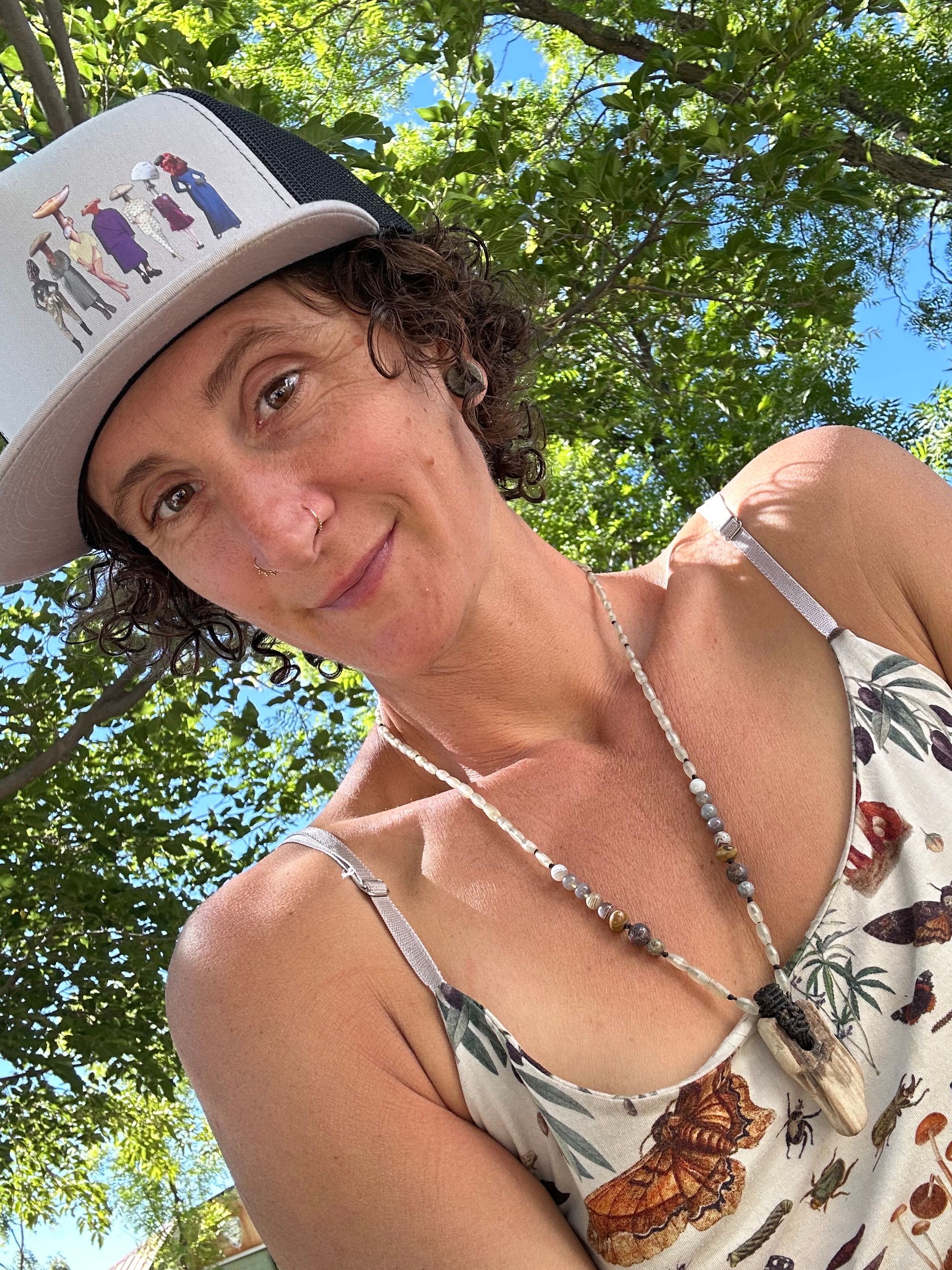 A woman wearing a trucker cap with images of mushroom head ladies on it where is a necklace of petrified wood with a shirt covered with images of mushrooms, butterflies, and plants