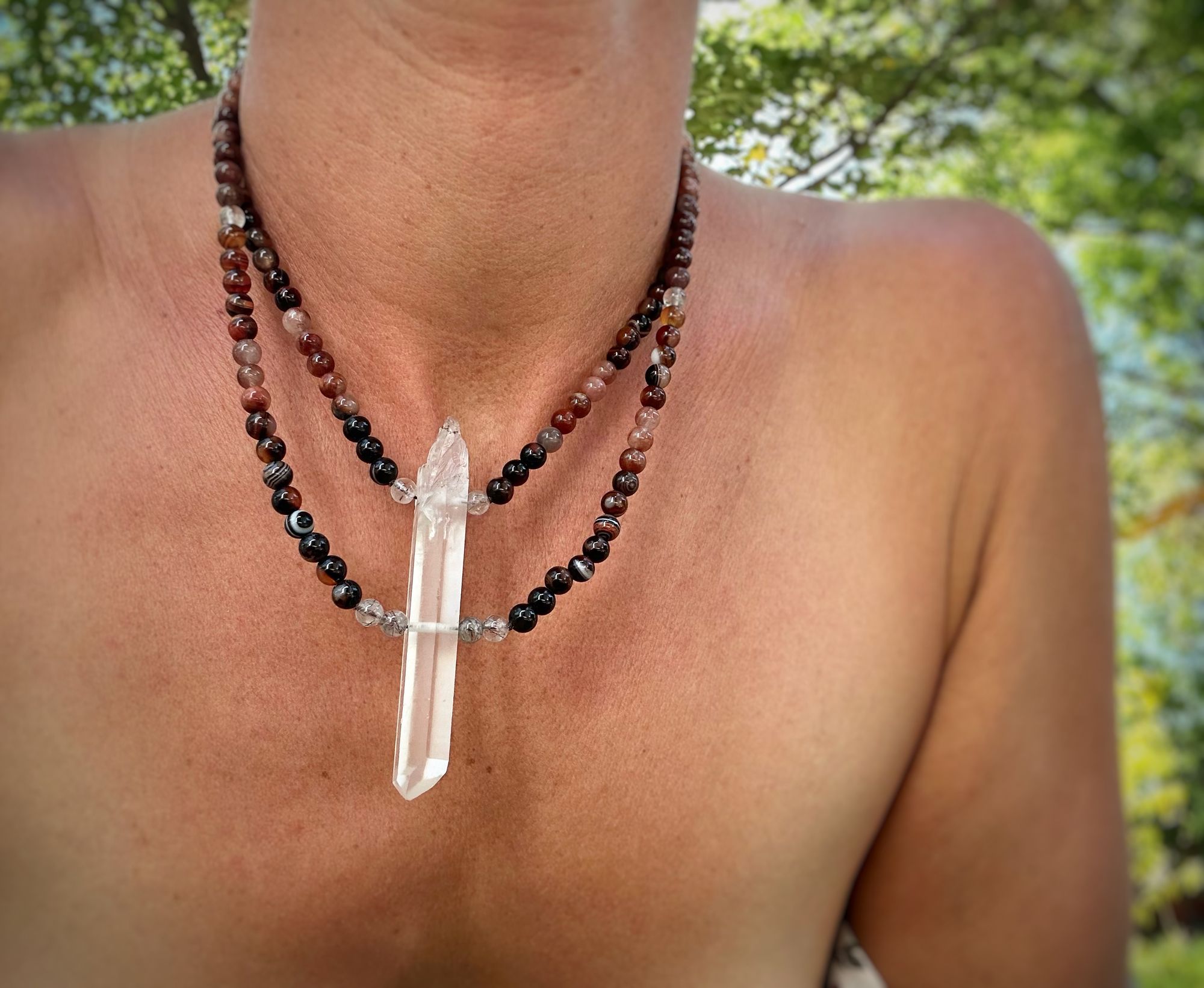 A woman wears a necklace of A necklace of clear quartz point with beads of deep hues of red ocre and soft pink