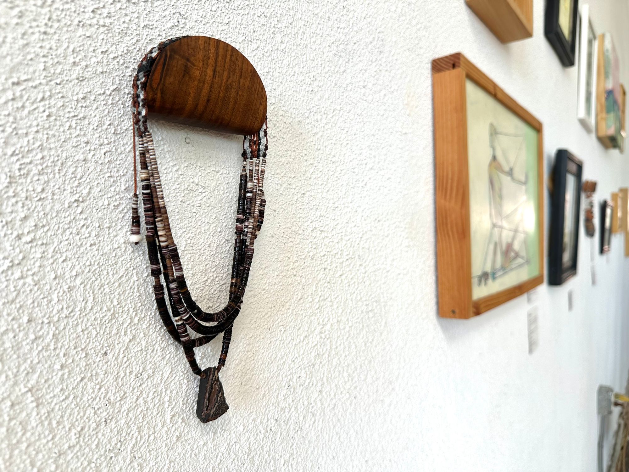 A necklace of white, cream, brown, and deep, rich purple shell beads With a mysterious Triangular brown and black stone with detailed basket, like knot work at the back Hings on a wooden hanger on a white wall