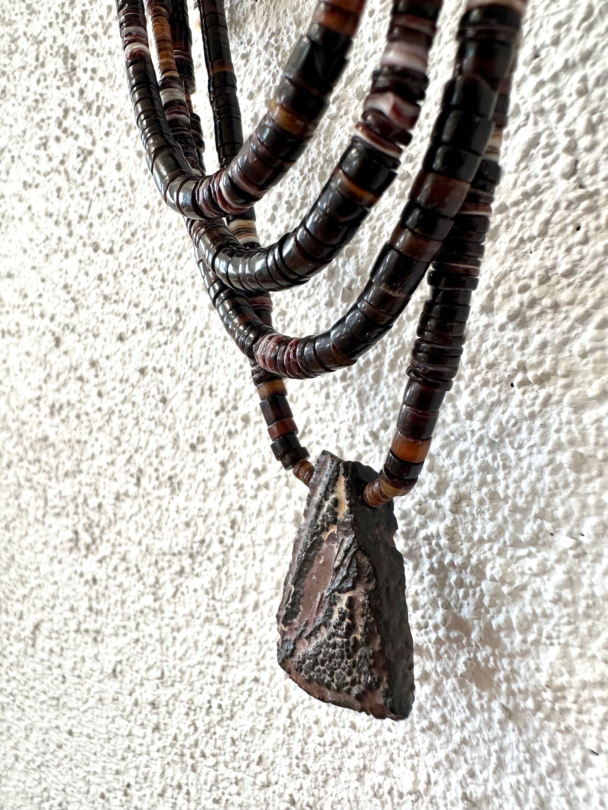 A necklace of white, cream, brown, and deep, rich purple shell beads With a mysterious Triangular brown and black stone with detailed basket, like knot work at the back Hings on a wooden hanger on a white wall