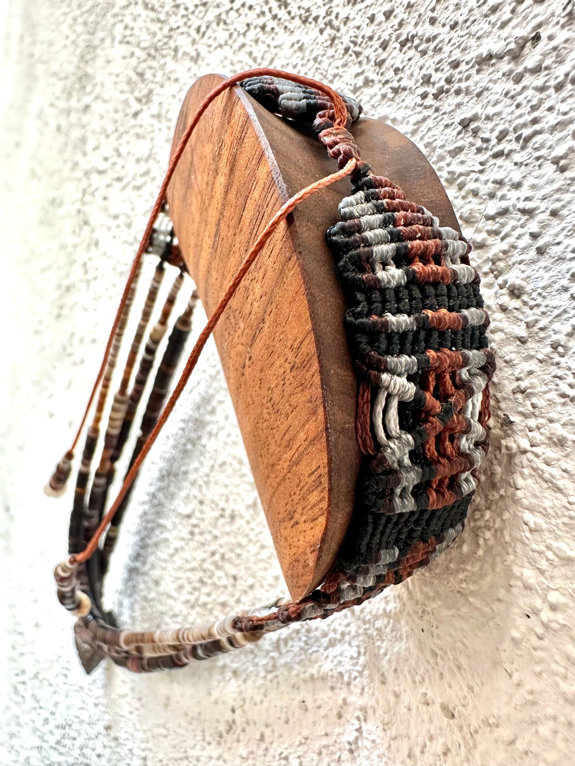 A necklace of white, cream, brown, and deep, rich purple shell beads With a mysterious Triangular brown and black stone with detailed basket, like knot work at the back Hings on a wooden hanger on a white wall