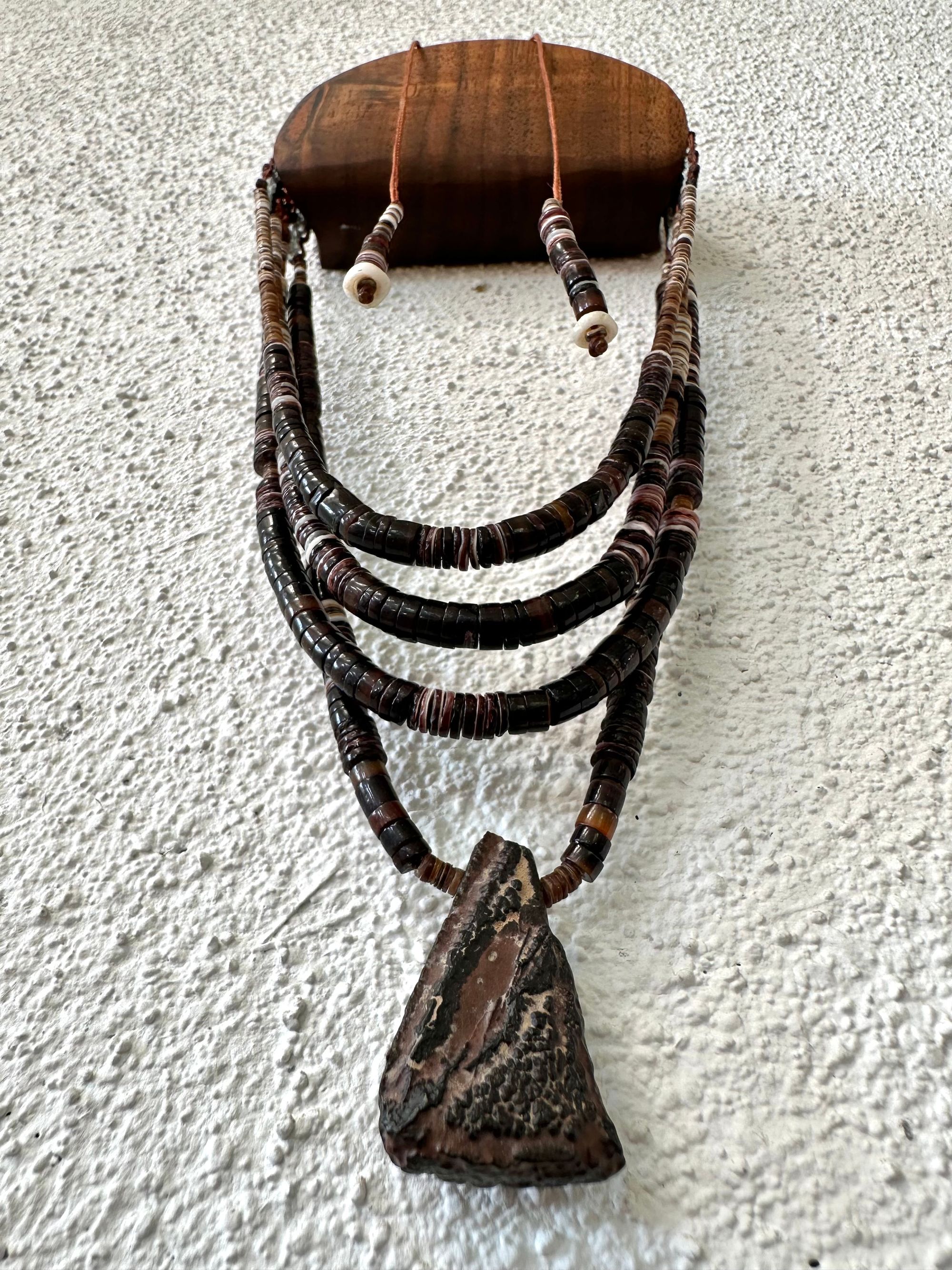 A necklace of white, cream, brown, and deep, rich purple shell beads With a mysterious Triangular brown and black stone with detailed basket, like knot work at the back Hings on a wooden hanger on a white wall