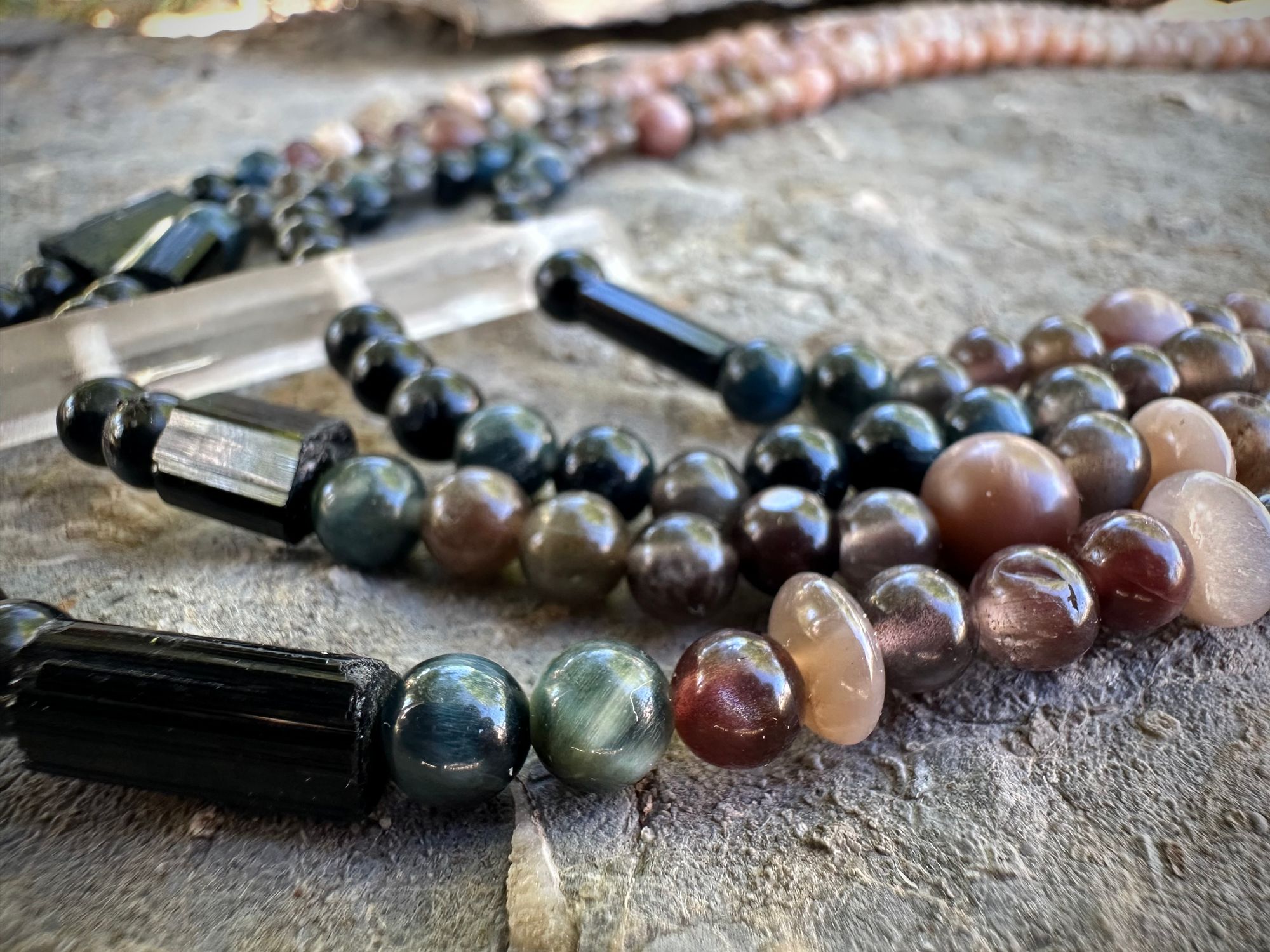 A necklace of black, deep, blue and dusty, pink, semi precious stone beads lays on a fossil filled stone