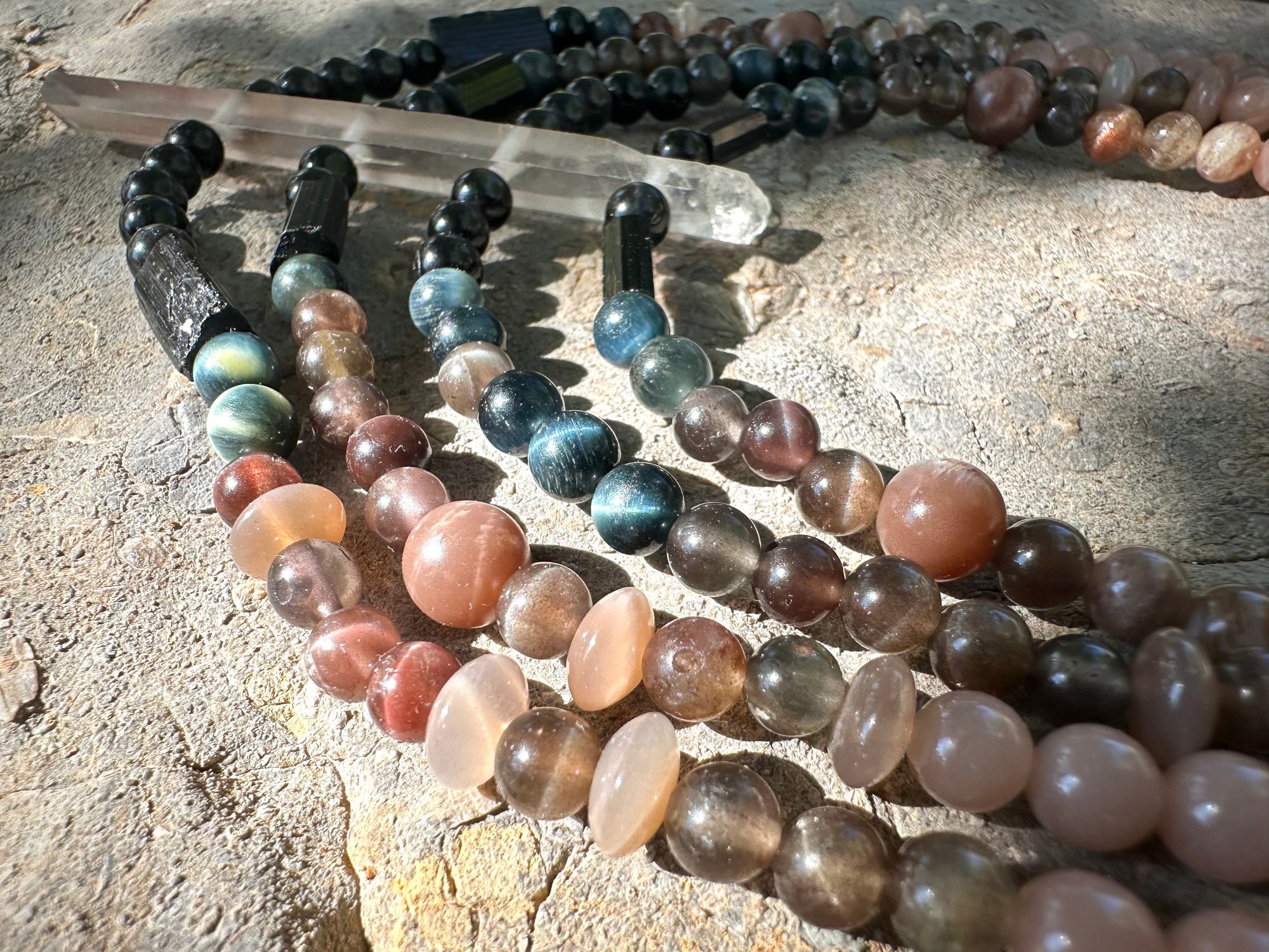 A necklace of black, deep, blue and dusty, pink, semi precious stone beads lays on a fossil filled stone