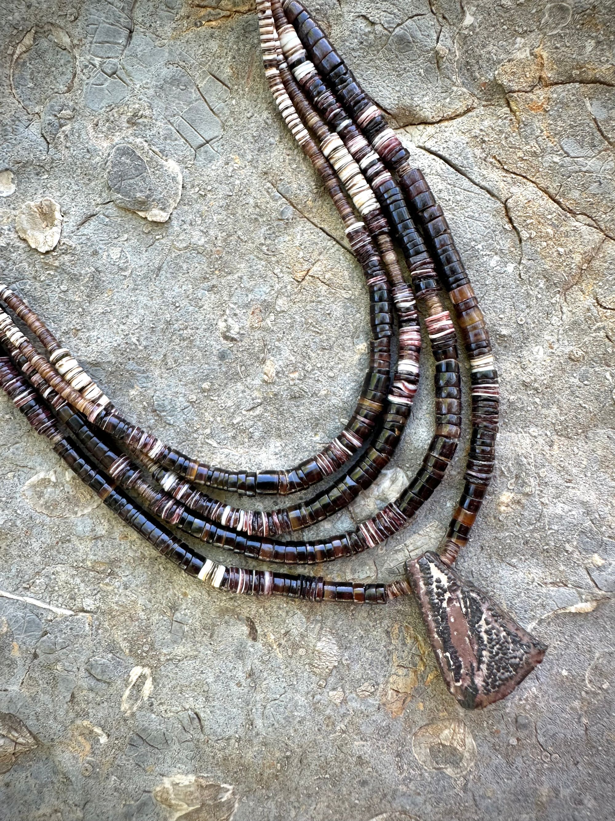 A necklace of white, cream, brown, and deep, rich purple shell beads With a mysterious Triangular brown and black stone with detailed basket, like knot work at the back lies on a Greystone filled with fossils