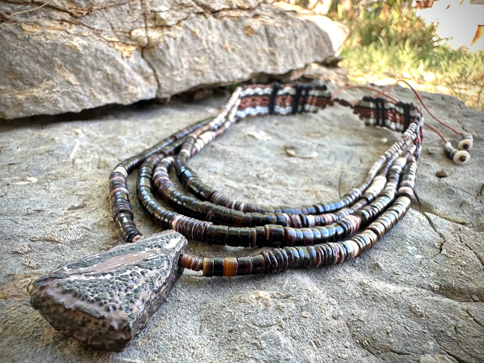 A necklace of white, cream, brown, and deep, rich purple shell beads With a mysterious Triangular brown and black stone with detailed basket, like knot work at the back lies on a Greystone filled with fossils