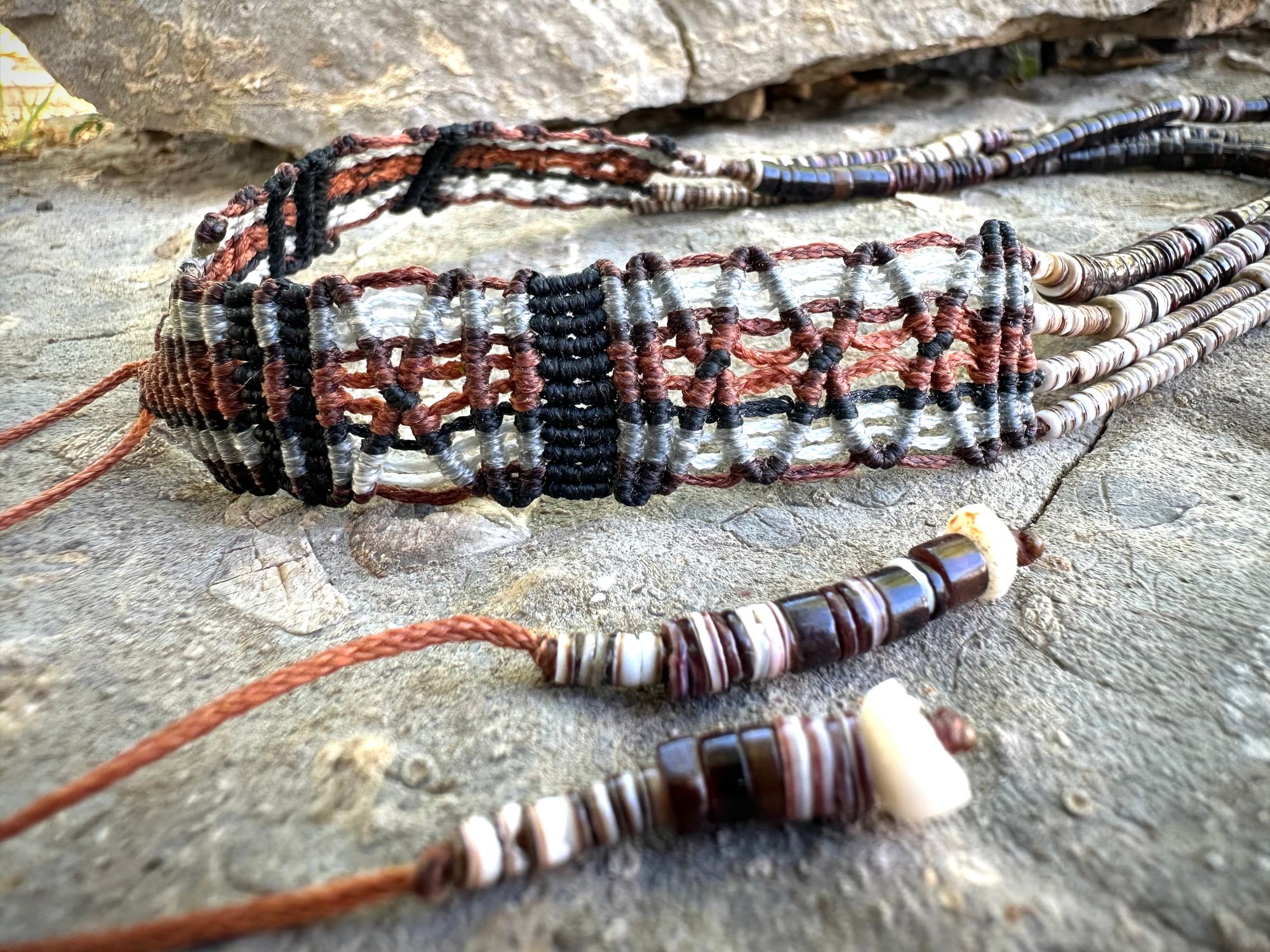 A necklace of white, cream, brown, and deep, rich purple shell beads With a mysterious Triangular brown and black stone with detailed basket, like knot work at the back lies on a Greystone filled with fossils