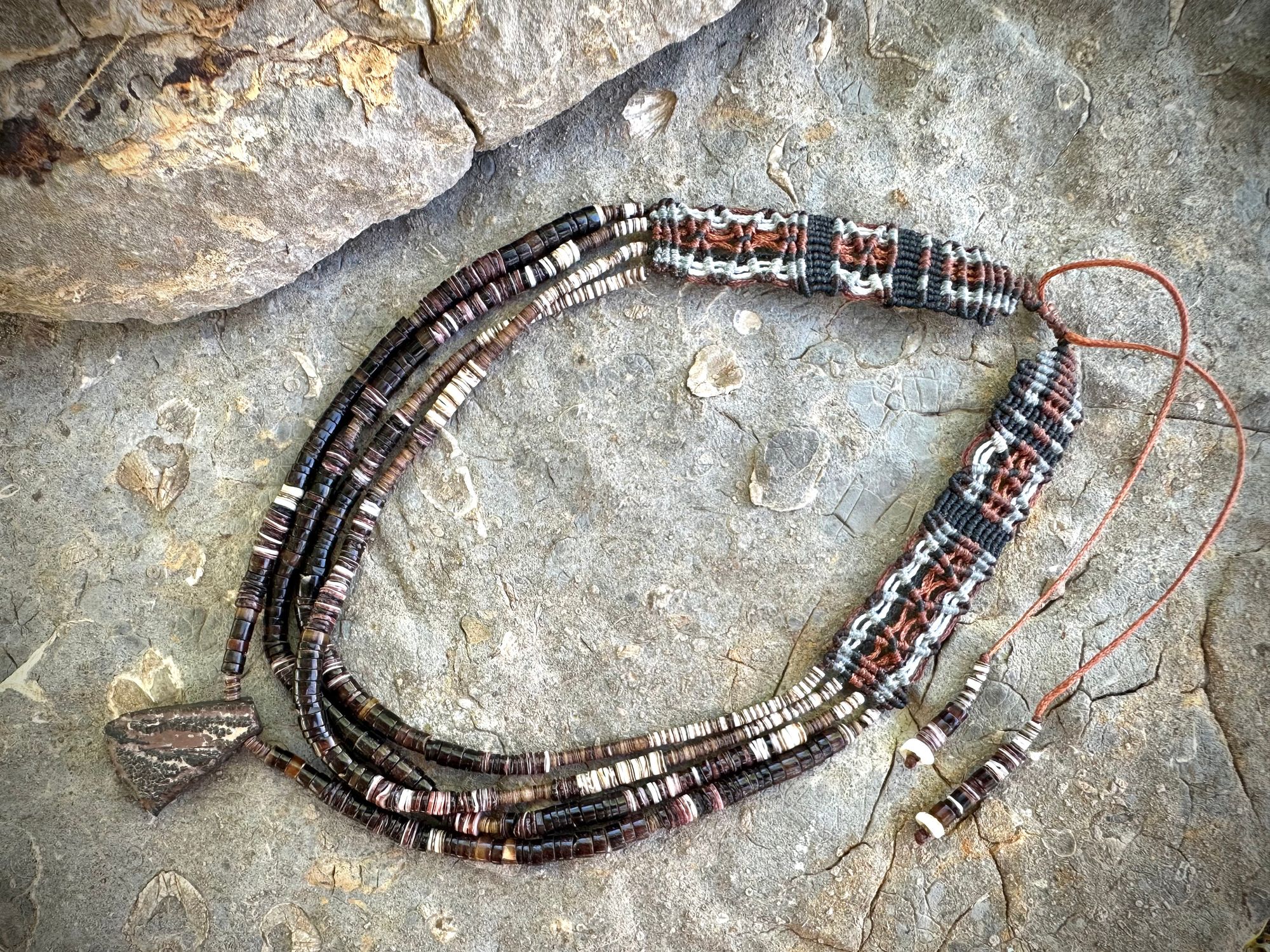 A necklace of white, cream, brown, and deep, rich purple shell beads With a mysterious Triangular brown and black stone with detailed basket, like knot work at the back lies on a Greystone filled with fossils