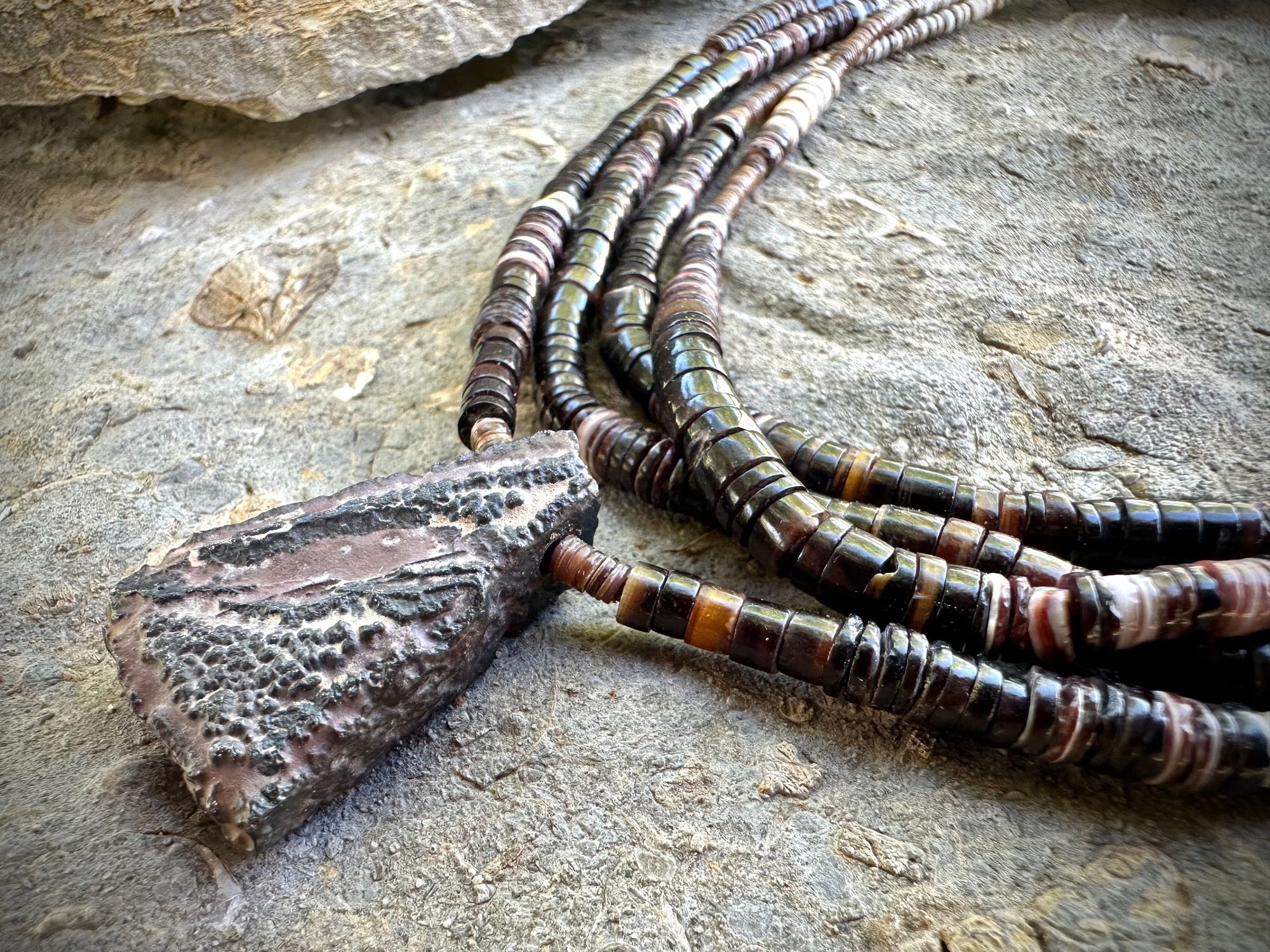 A necklace of white, cream, brown, and deep, rich purple shell beads With a mysterious Triangular brown and black stone with detailed basket, like knot work at the back lies on a Greystone filled with fossils