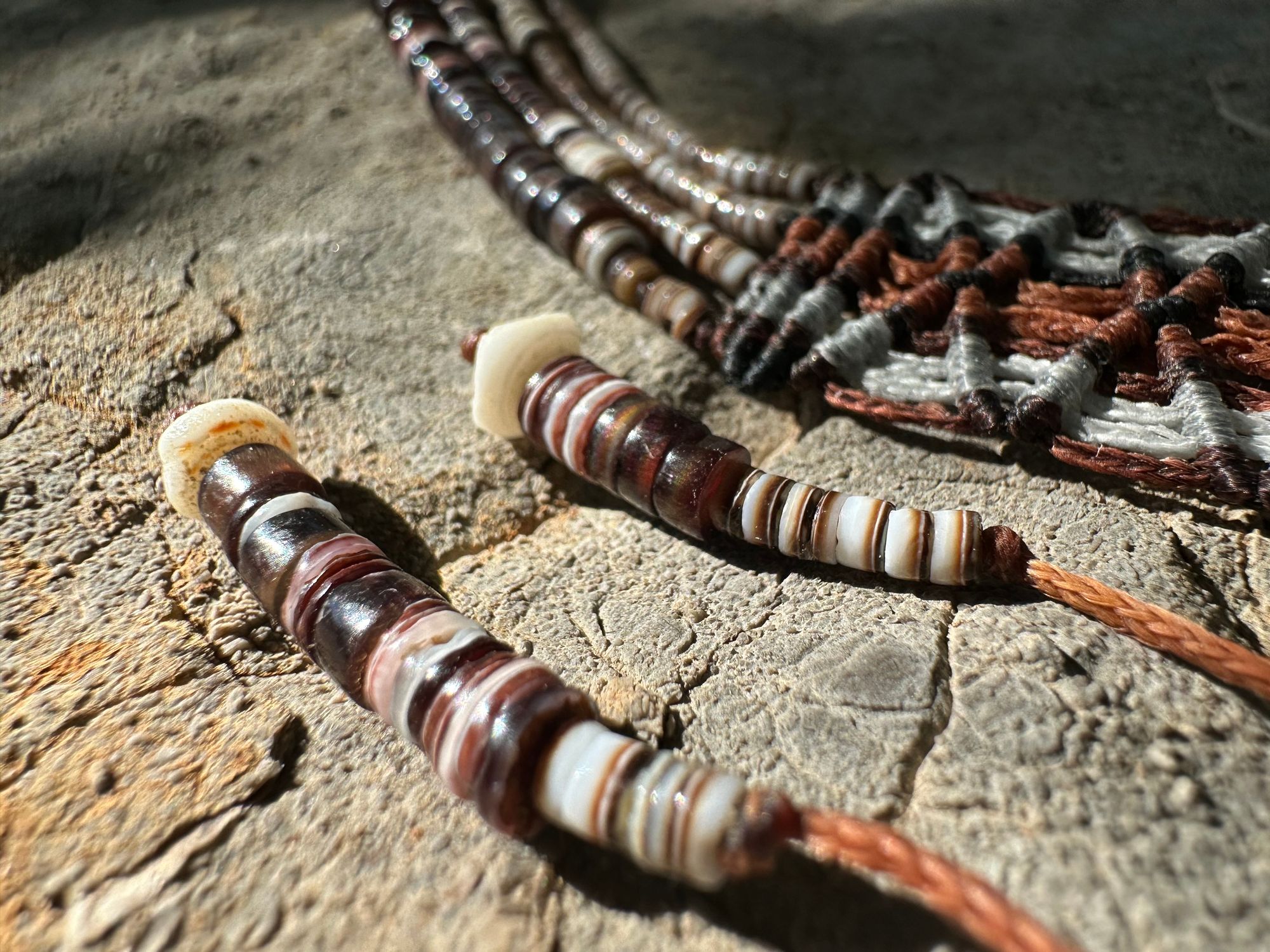 A necklace of white, cream, brown, and deep, rich purple shell beads With a mysterious Triangular brown and black stone with detailed basket, like knot work at the back lies on a Greystone filled with fossils