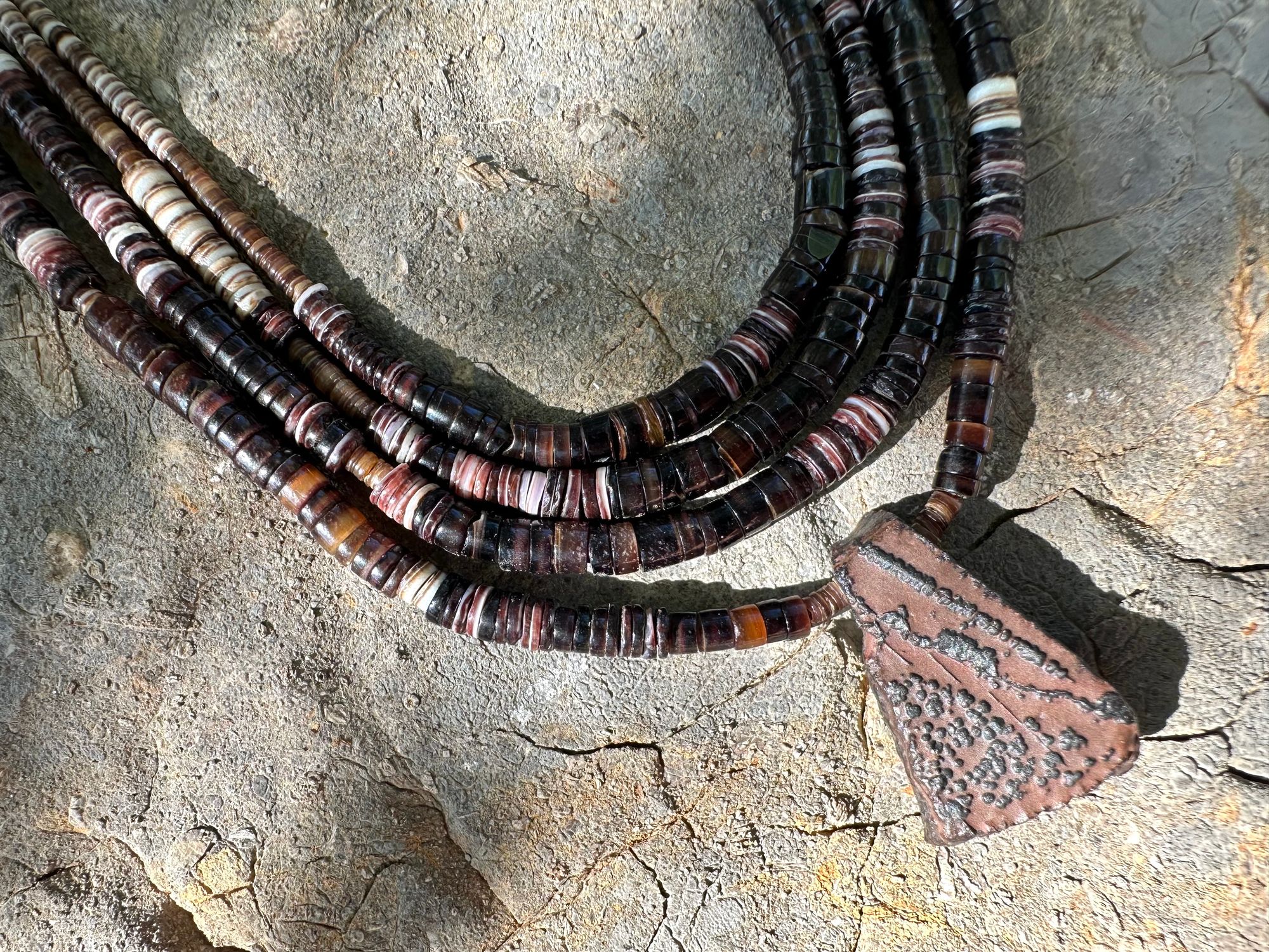 A necklace of white, cream, brown, and deep, rich purple shell beads With a mysterious Triangular brown and black stone with detailed basket, like knot work at the back lies on a Greystone filled with fossils