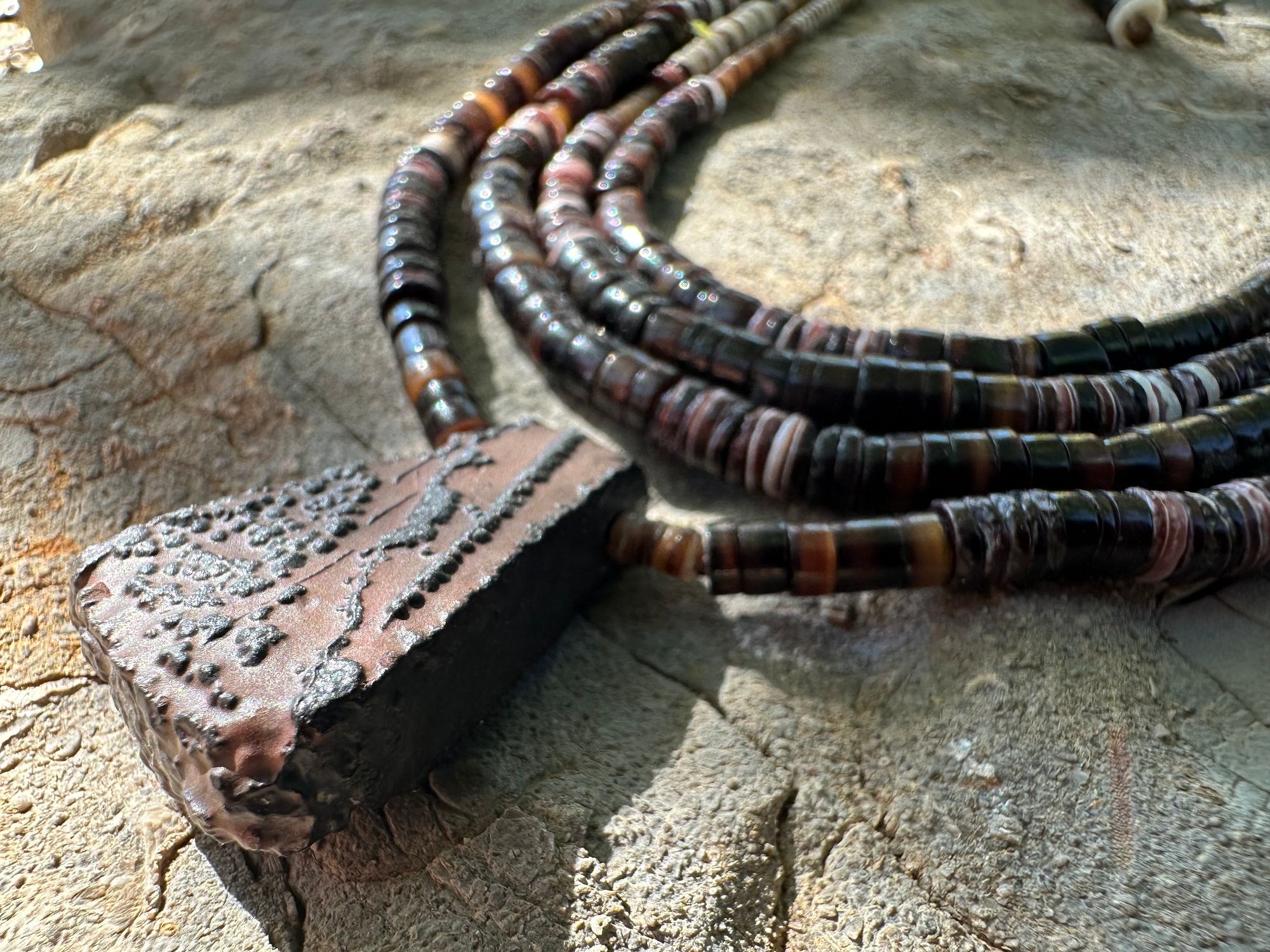 A necklace of white, cream, brown, and deep, rich purple shell beads With a mysterious Triangular brown and black stone with detailed basket, like knot work at the back lies on a Greystone filled with fossils