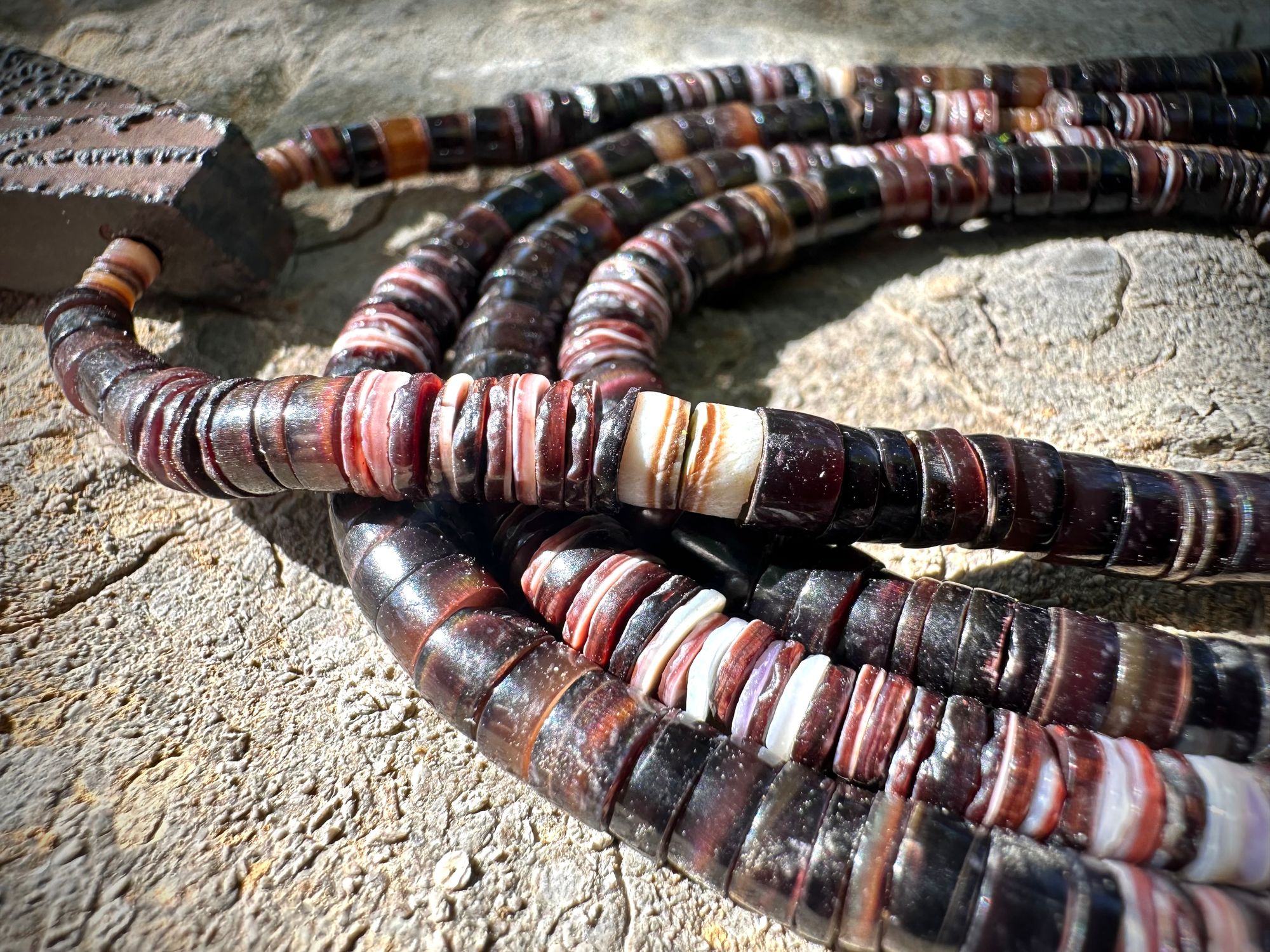 A necklace of white, cream, brown, and deep, rich purple shell beads With a mysterious Triangular brown and black stone with detailed basket, like knot work at the back lies on a Greystone filled with fossils