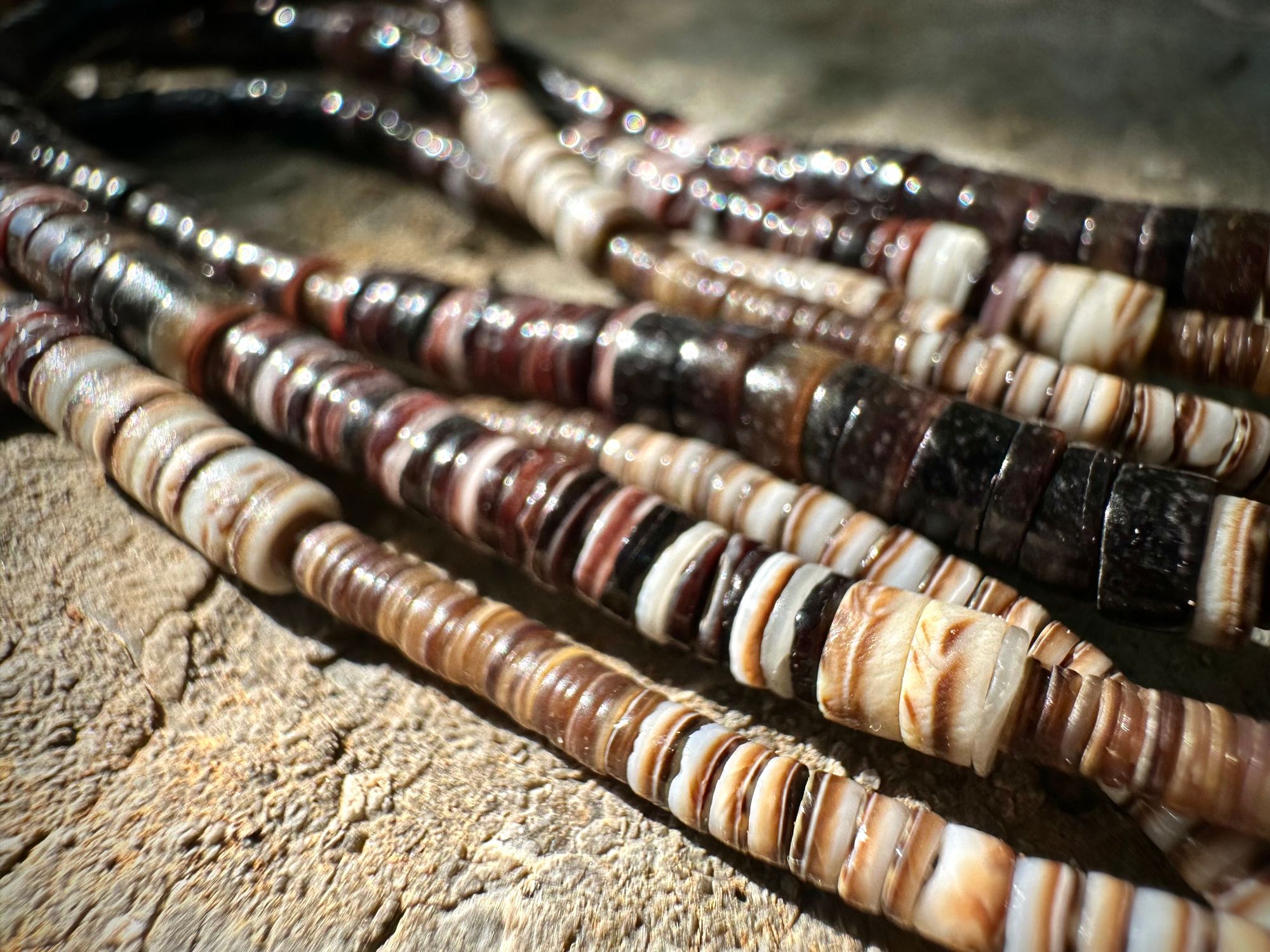 A necklace of white, cream, brown, and deep, rich purple shell beads With a mysterious Triangular brown and black stone with detailed basket, like knot work at the back lies on a Greystone filled with fossils