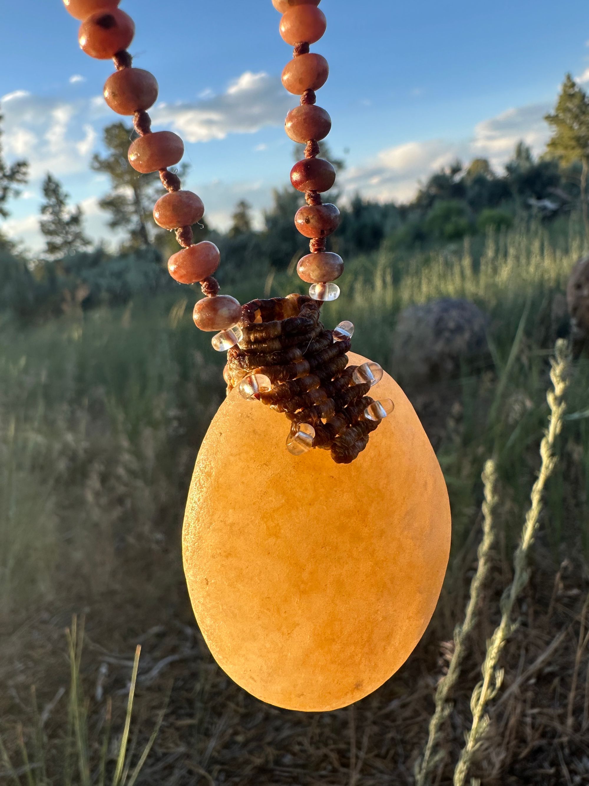 An ocean tumble stone formed into a work and beaded necklace, glows in the light of sunset a grassy forest background