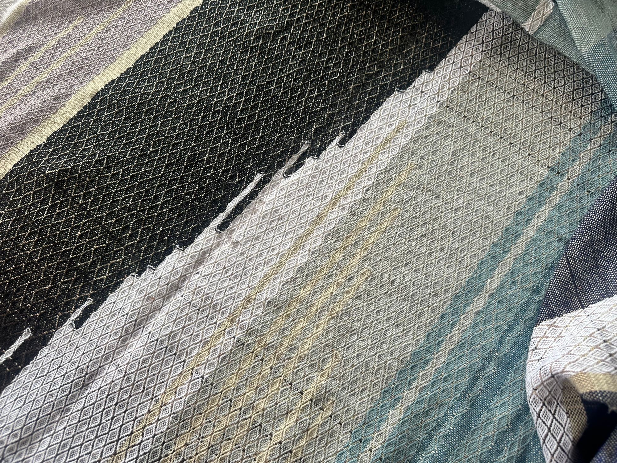 A piece of handwoven, diamond patterned, with wave-like designs fabric in shades of white, to blue, to black lays on a white floor.