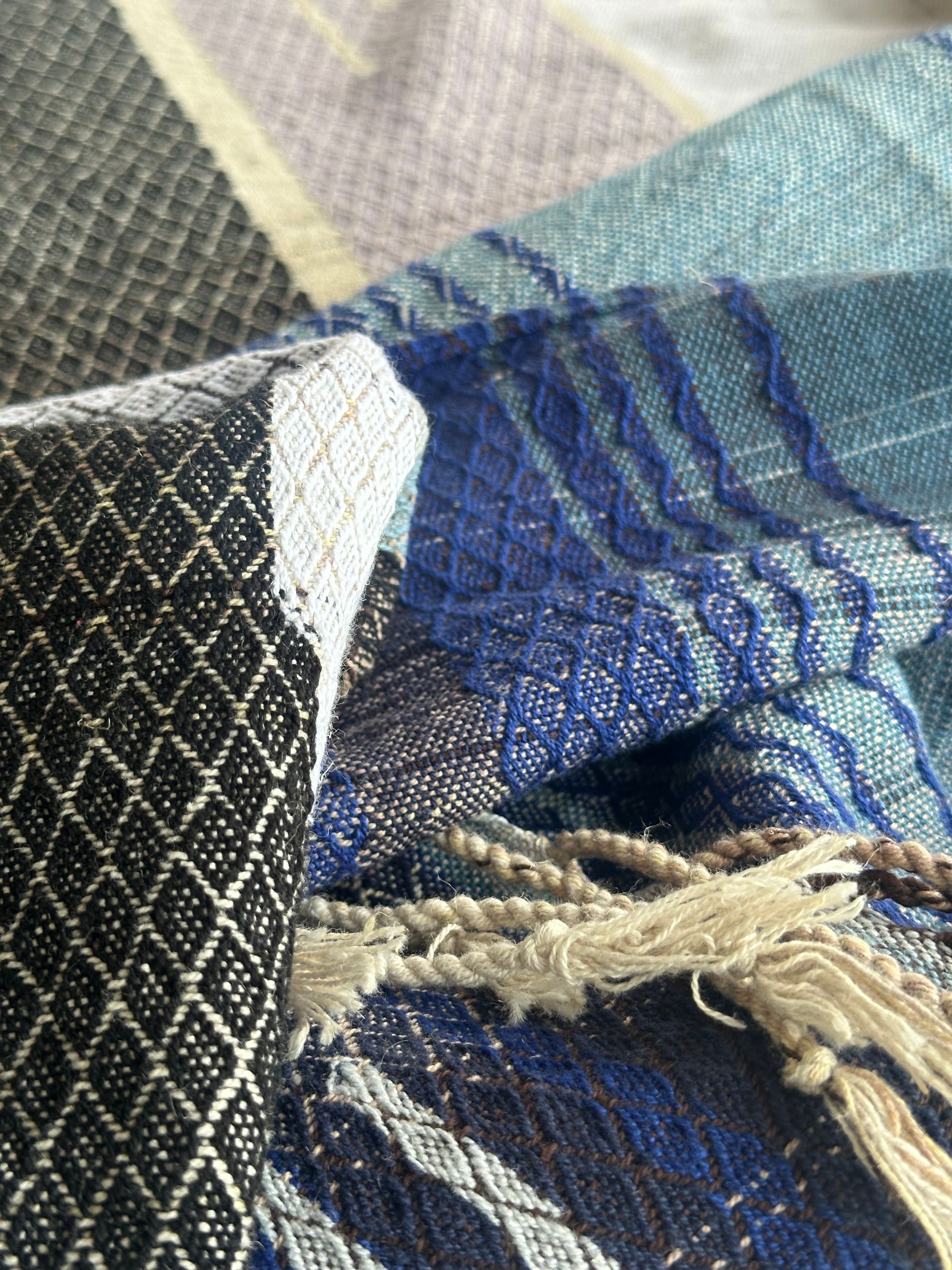 A piece of handwoven, diamond patterned, with wave-like designs fabric in shades of white, to blue, to black lays on a white floor.