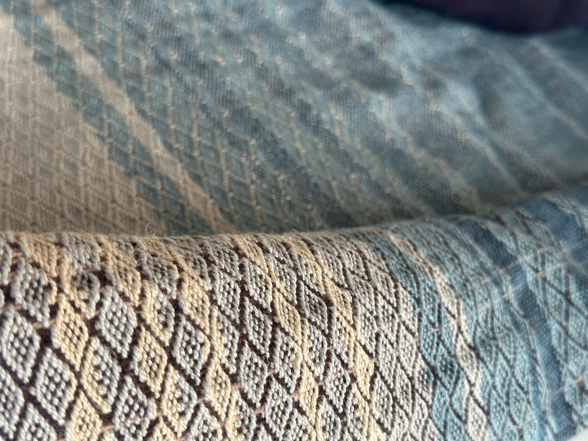 a detail of A piece of handwoven, diamond patterned, with wave-like designs fabric in shades of white, to blue, to black lays on a white floor.