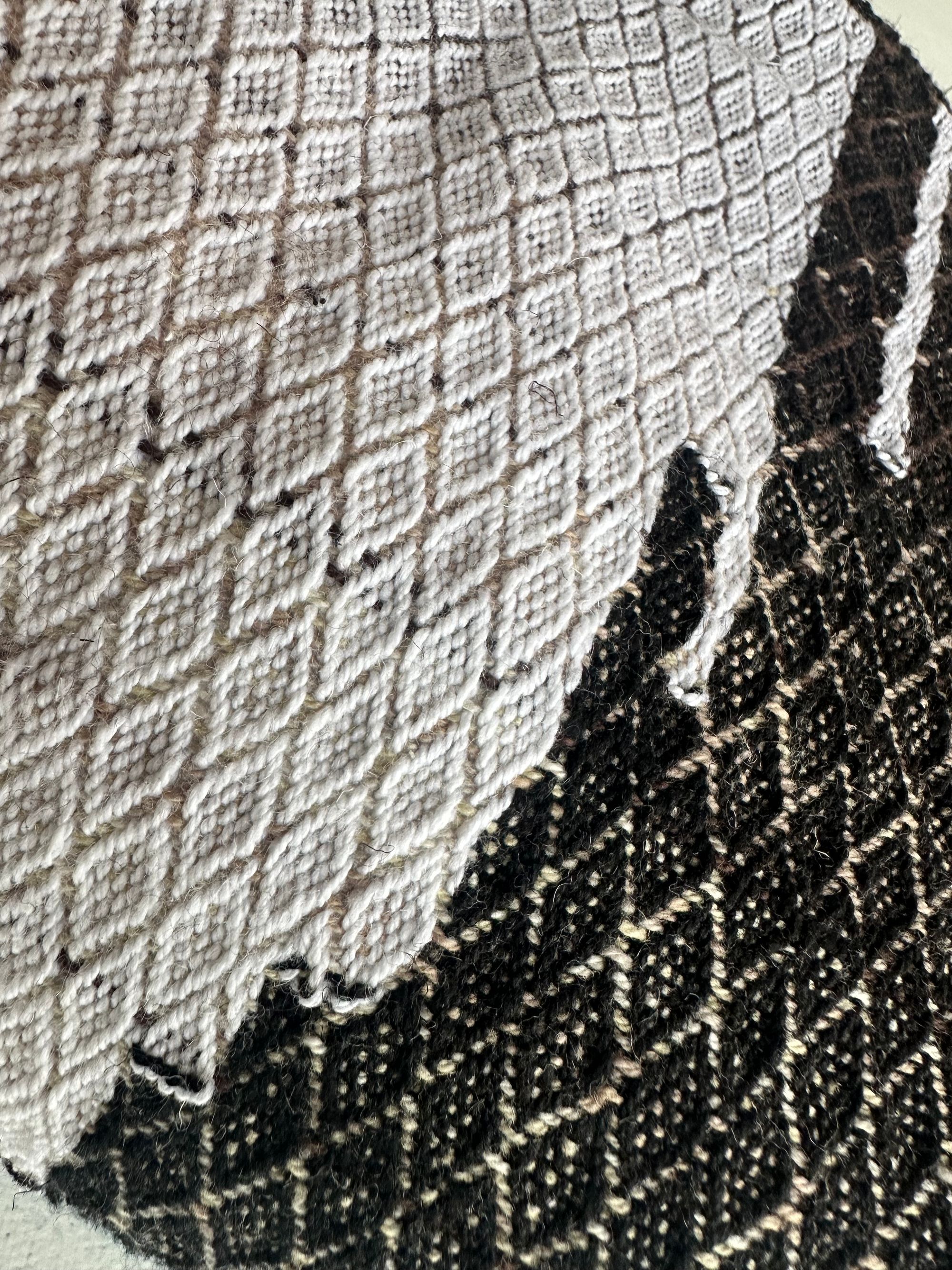 a detail of the black and white diamond pattern on A piece of handwoven, diamond patterned, with wave-like designs fabric in shades of white, to blue, to black lays on a white floor.