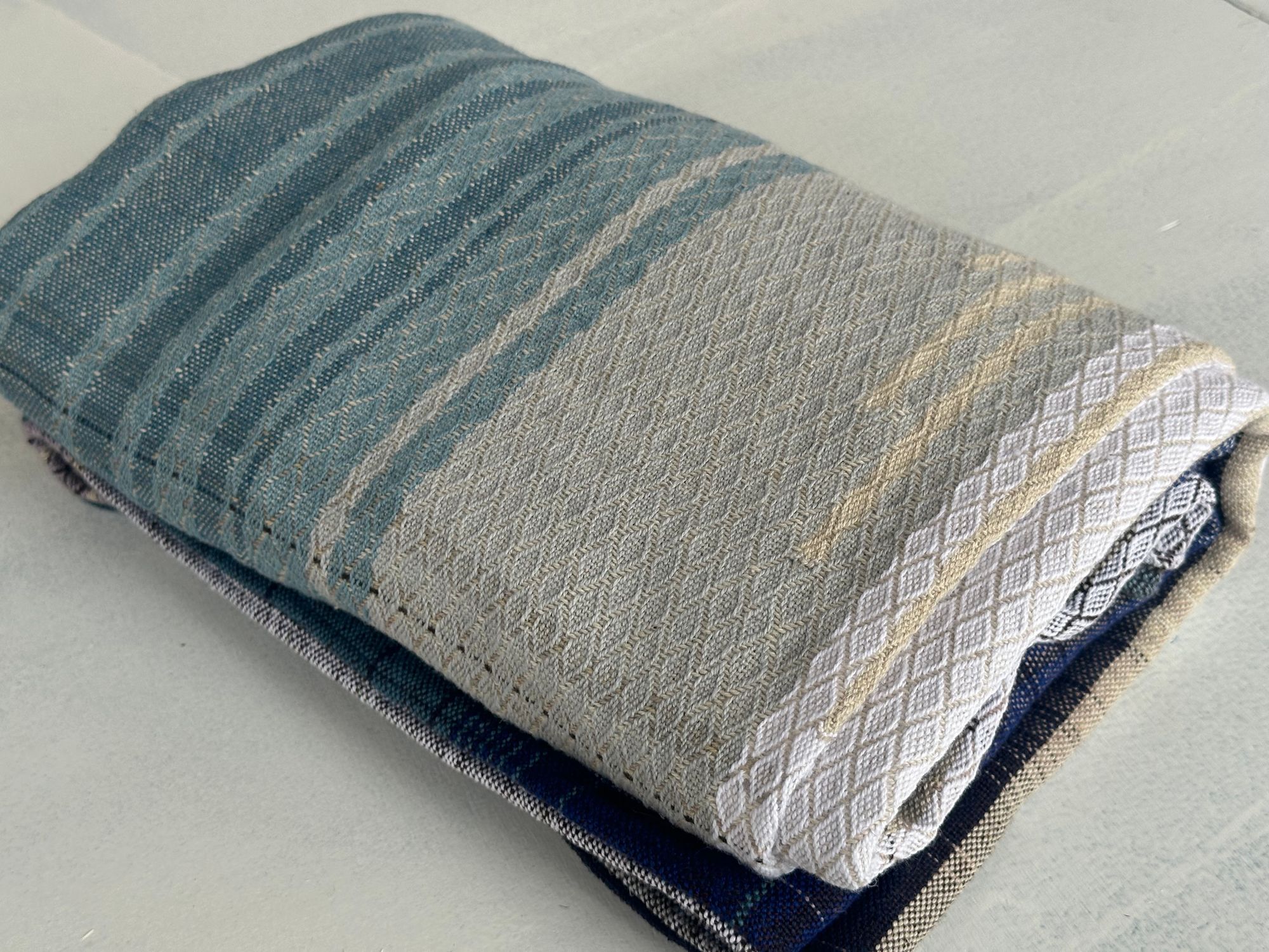 A piece of handwoven, diamond patterned, with wave-like designs fabric in shades of white, to blue, to black lays folded on a white floor.