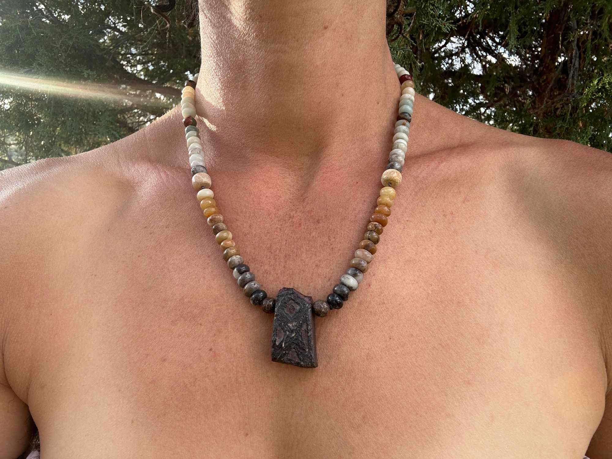 A woman wears A necklace made of Gray, black and brown tsesit, A while, ancient stone is formed into an necklace with a rainbow of Jasper bees, petrified, dinosaur bone and fossilized coral