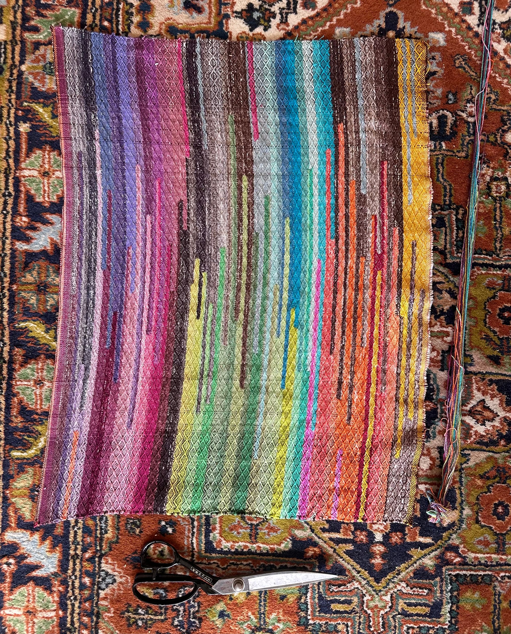 A small piece of hand woven fabric with a diamond texture, pattern, and bands of all the colors of the rainbow woven into it