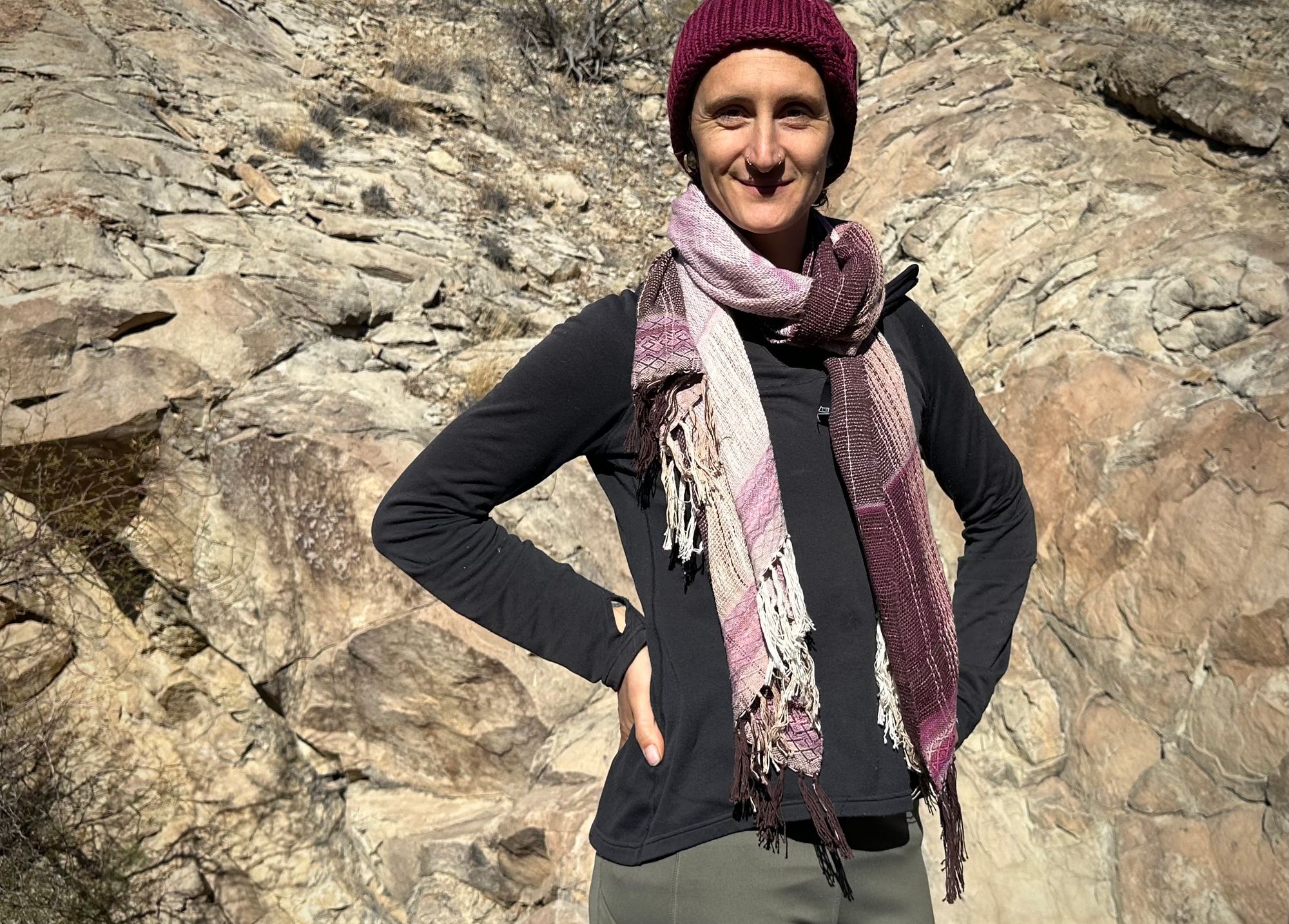 A woman stands in front of a cliff with a pink naturally dyed scarf