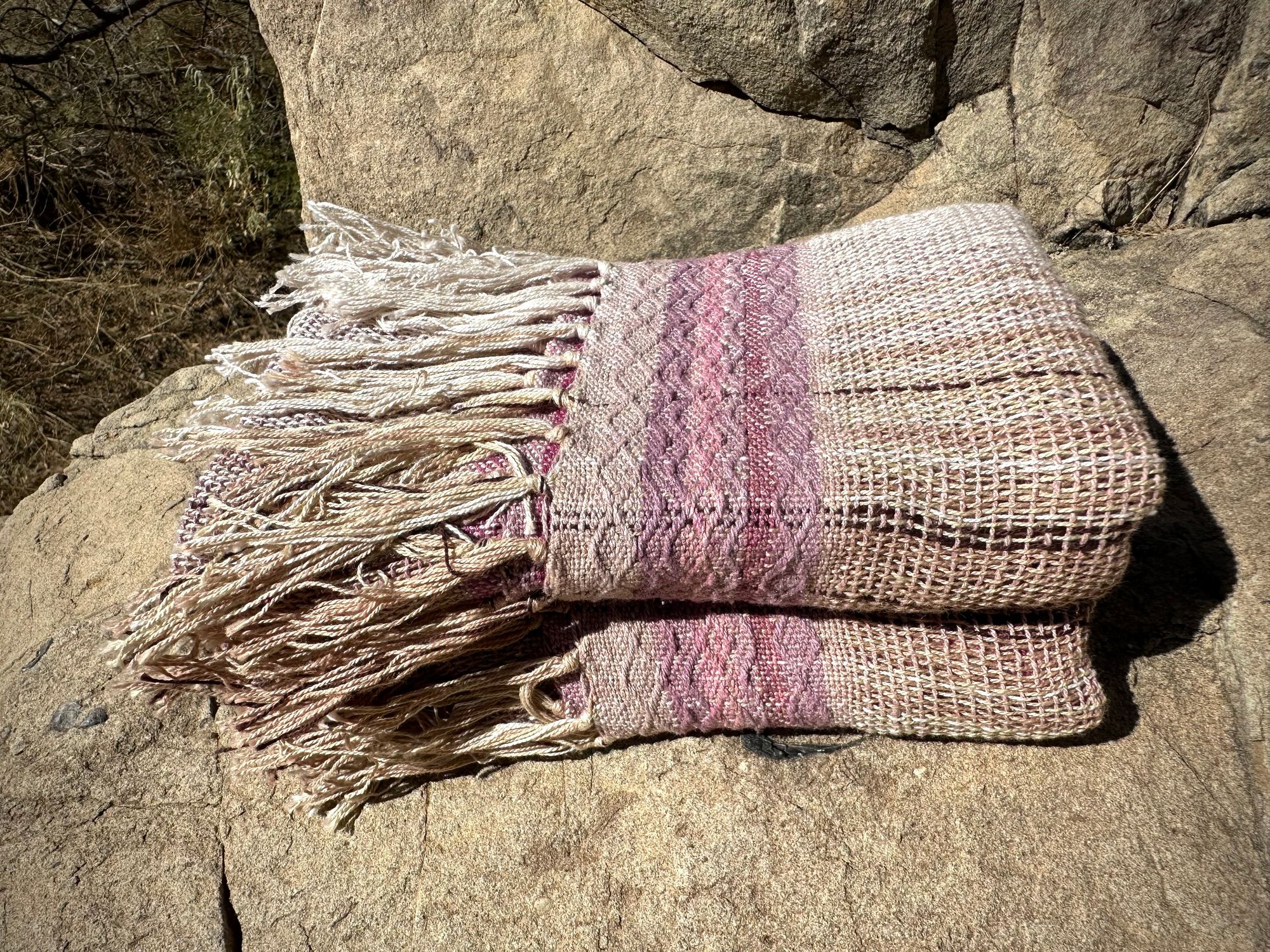 A naturally dyed scarf in pinks and lavender lays folded on a gray cliff