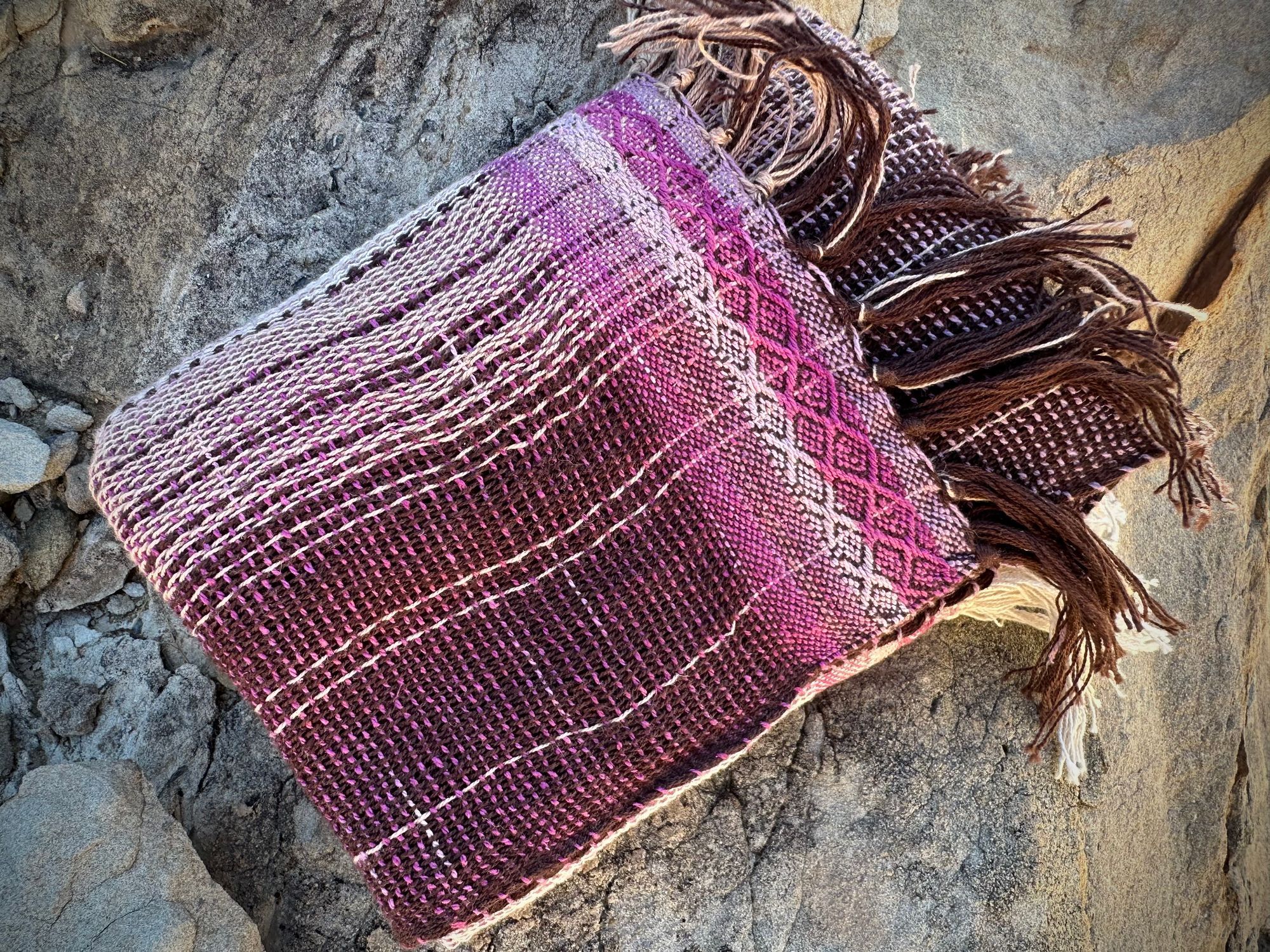 A naturally dyed scarf in pinks and lavender lays folded on a gray cliff