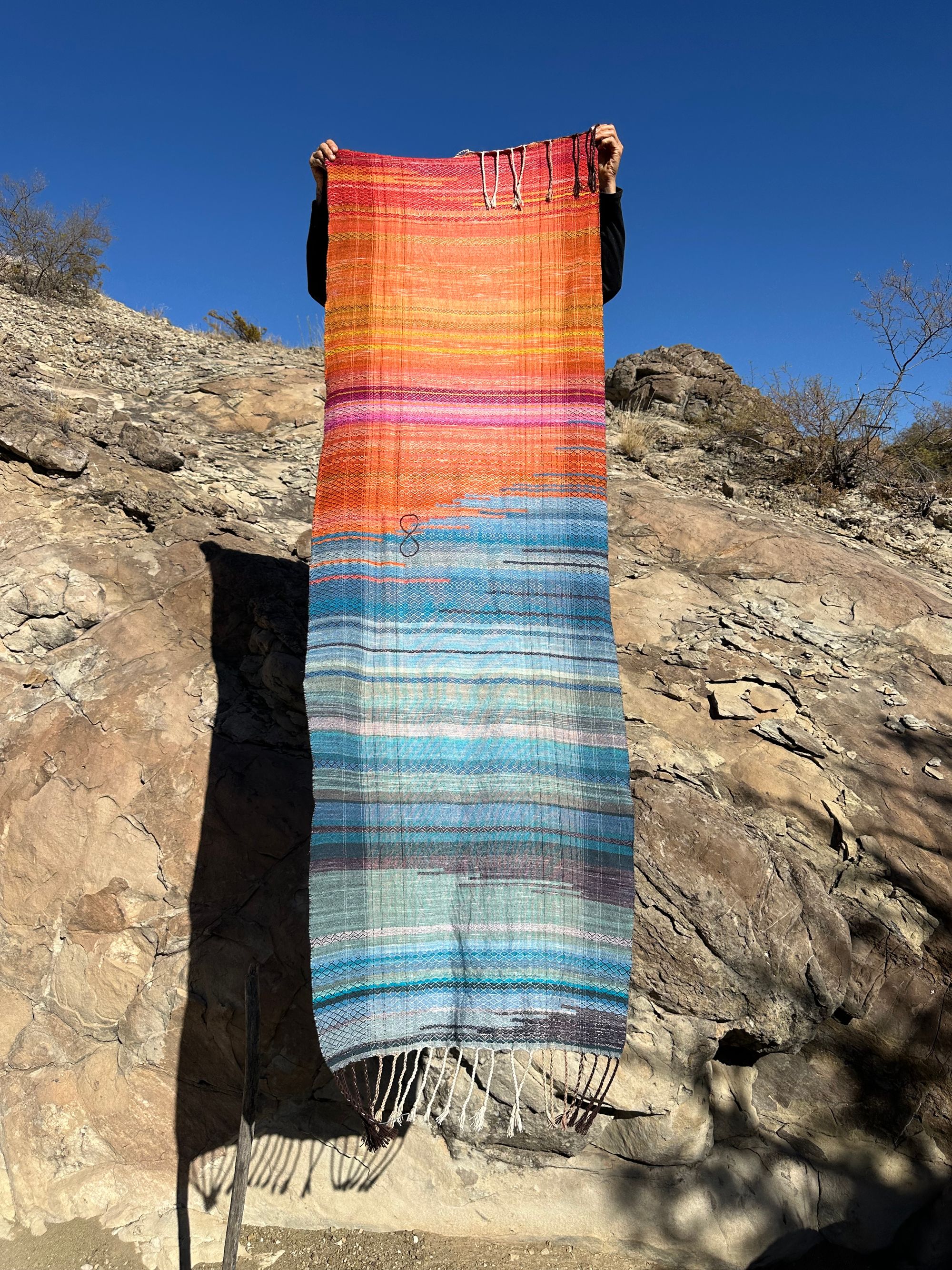 A woman holds A handwoven shawl, half the colors of fire and half the colors of ice as she stands on a cliff it has a brown infinity sign in the middle of it