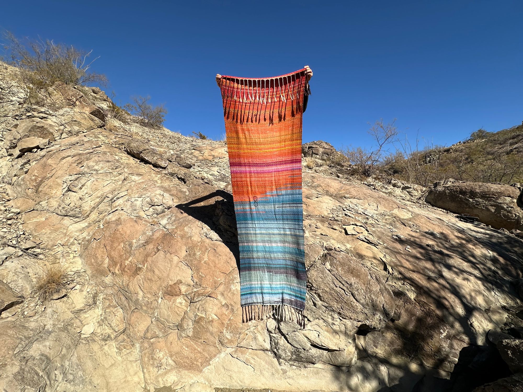 A woman holds A handwoven shawl, half the colors of fire and half the colors of ice as she stands on a cliff it has a brown infinity sign in the middle of it