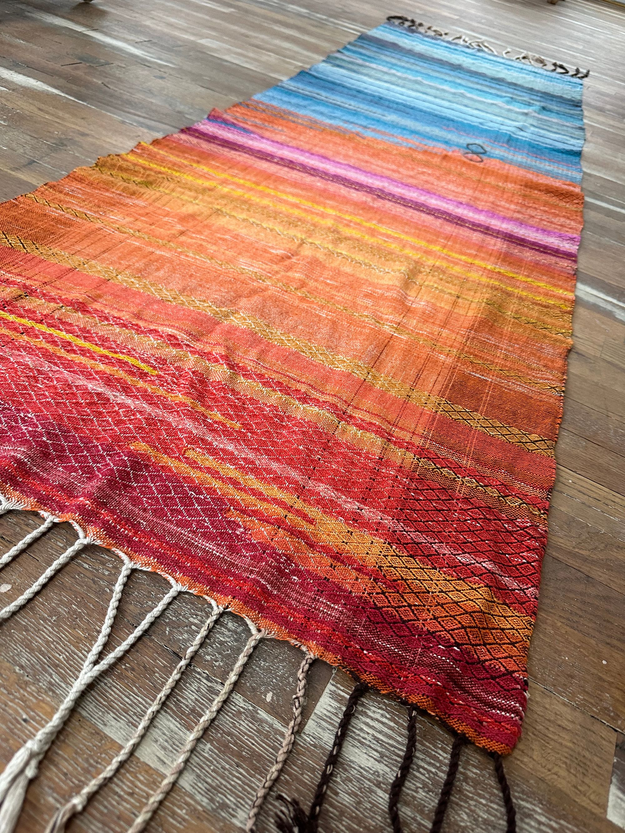 A handwoven shawl, half the colors of fire and half the colors of ice lays on a wooden floor. it has a brown infinity sign in the middle of it