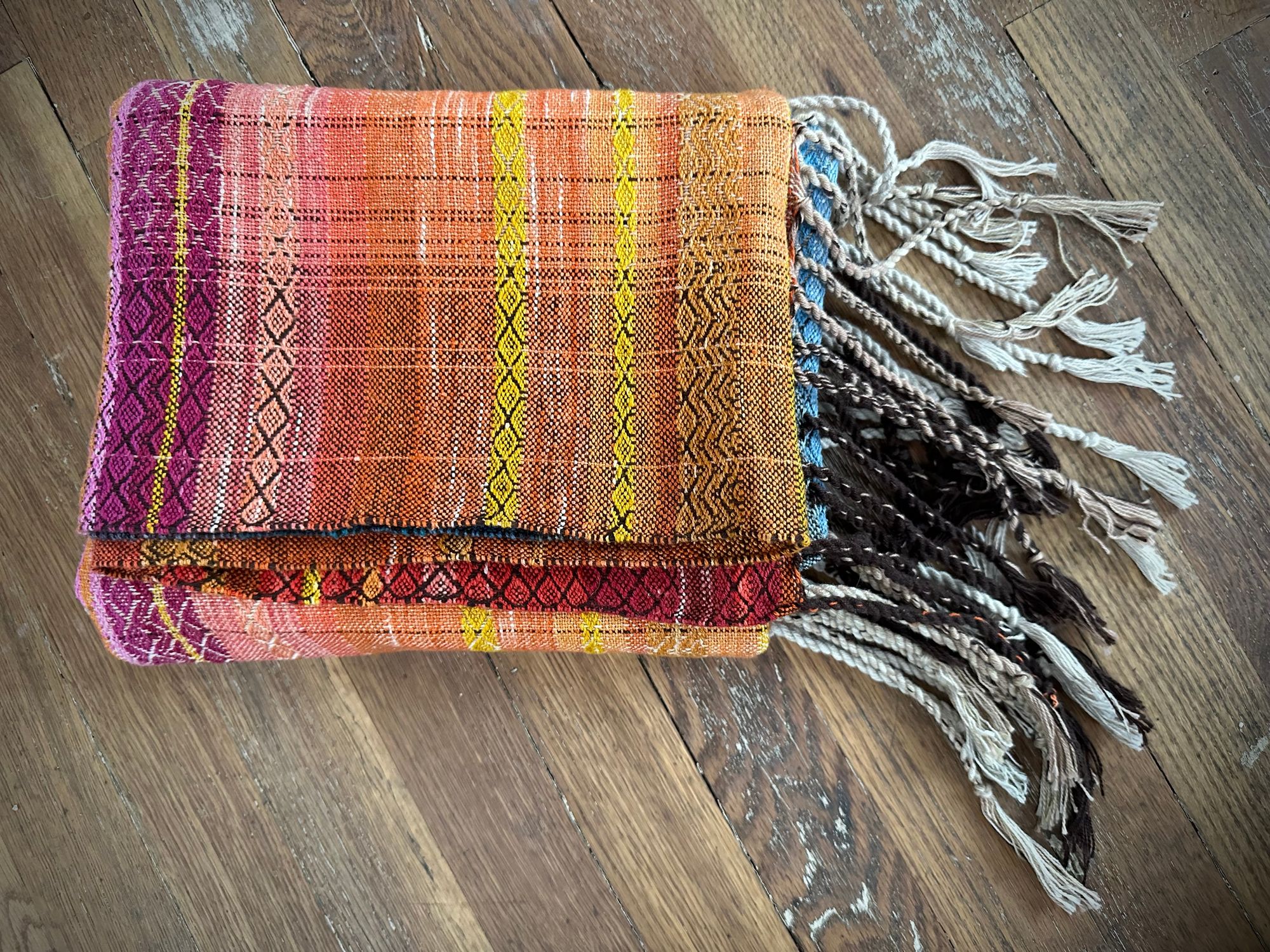 A handwoven shawl, half the colors of fire and half the colors of ice lays on a wooden floor. it has a brown infinity sign in the middle of it