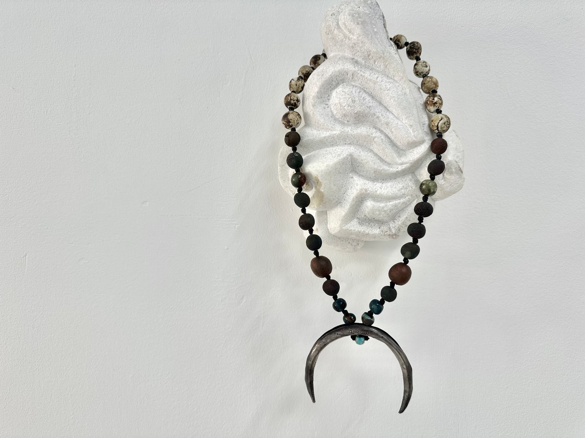 a necklace with a silver crescent moon and clay beads in earthy brown, blue and gray colors hangs from an abstract marble sculpture. The beads look like lava or stone