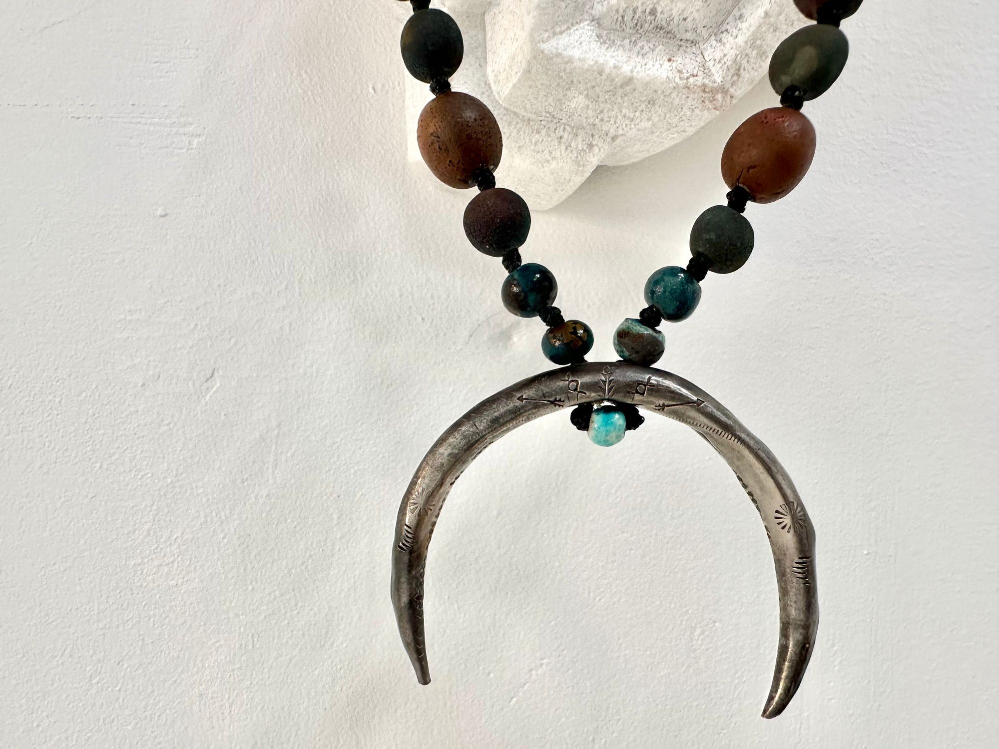 a necklace with a silver crescent moon and clay beads in earthy brown, blue and gray colors hangs from an abstract marble sculpture. The beads look like lava or stone