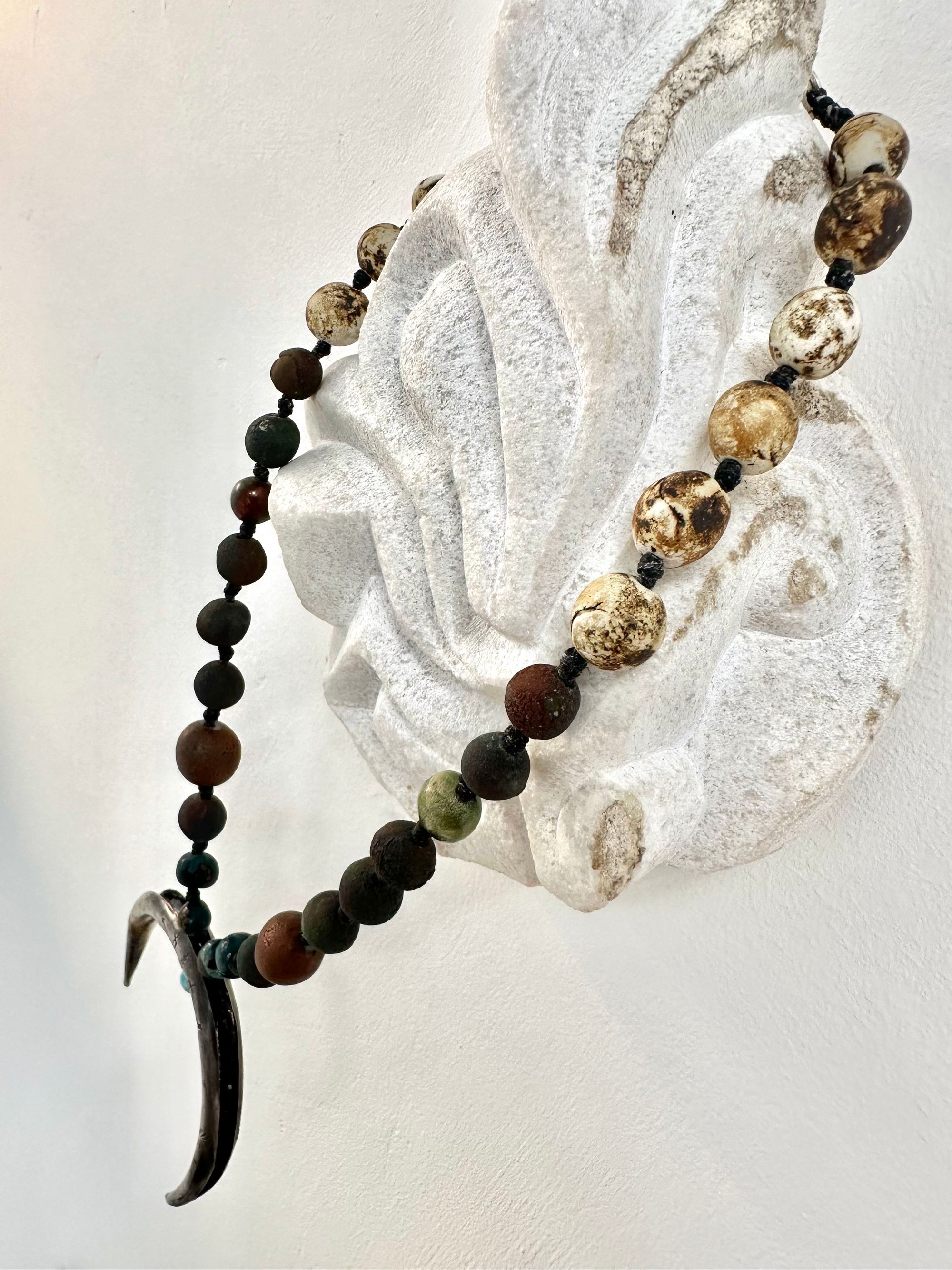 a necklace with a silver crescent moon and clay beads in earthy brown, blue and gray colors hangs from an abstract marble sculpture. The beads look like lava or stone