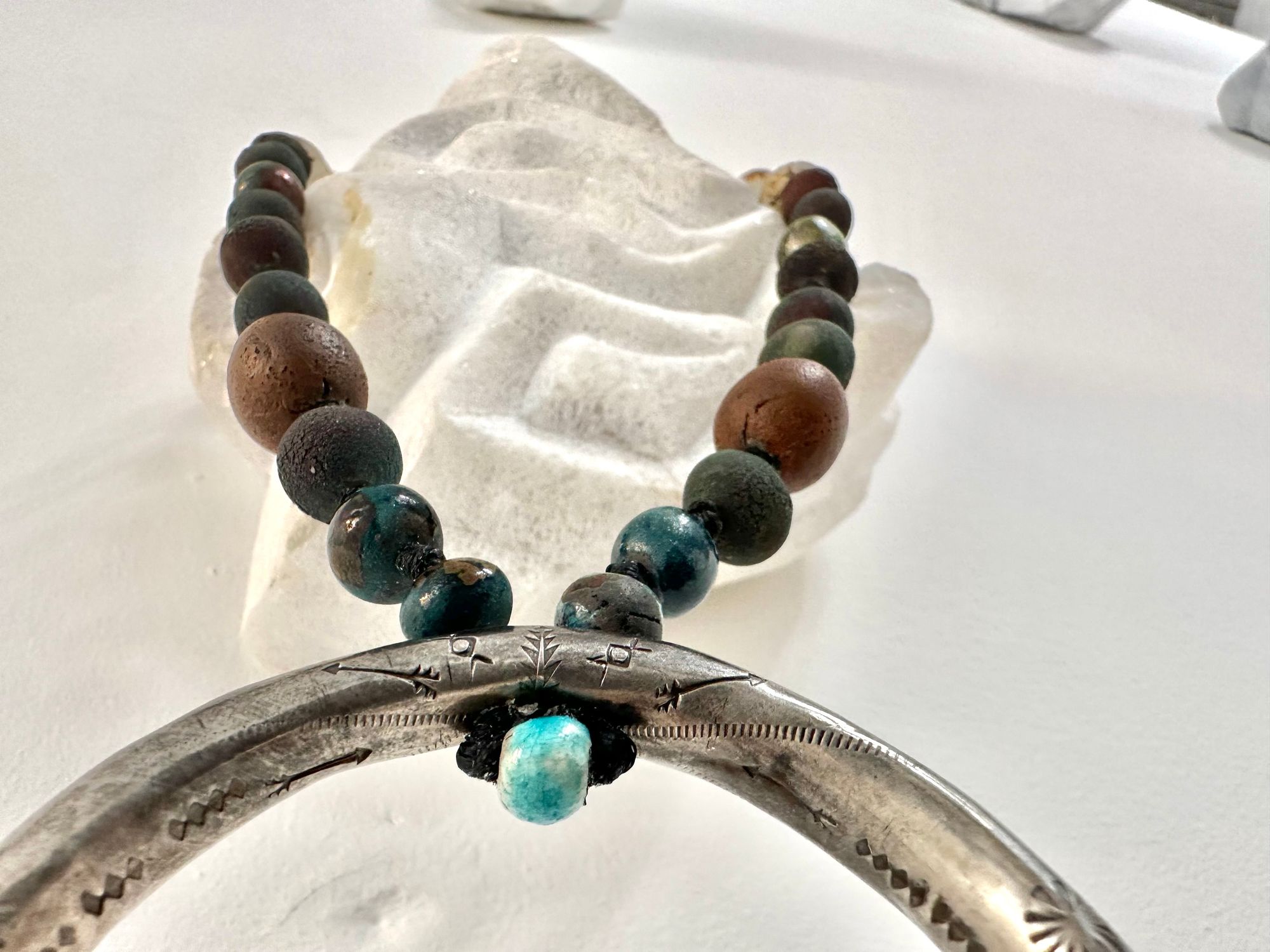 a necklace with a silver crescent moon and clay beads in earthy brown, blue and gray colors hangs from an abstract marble sculpture. The beads look like lava or stone