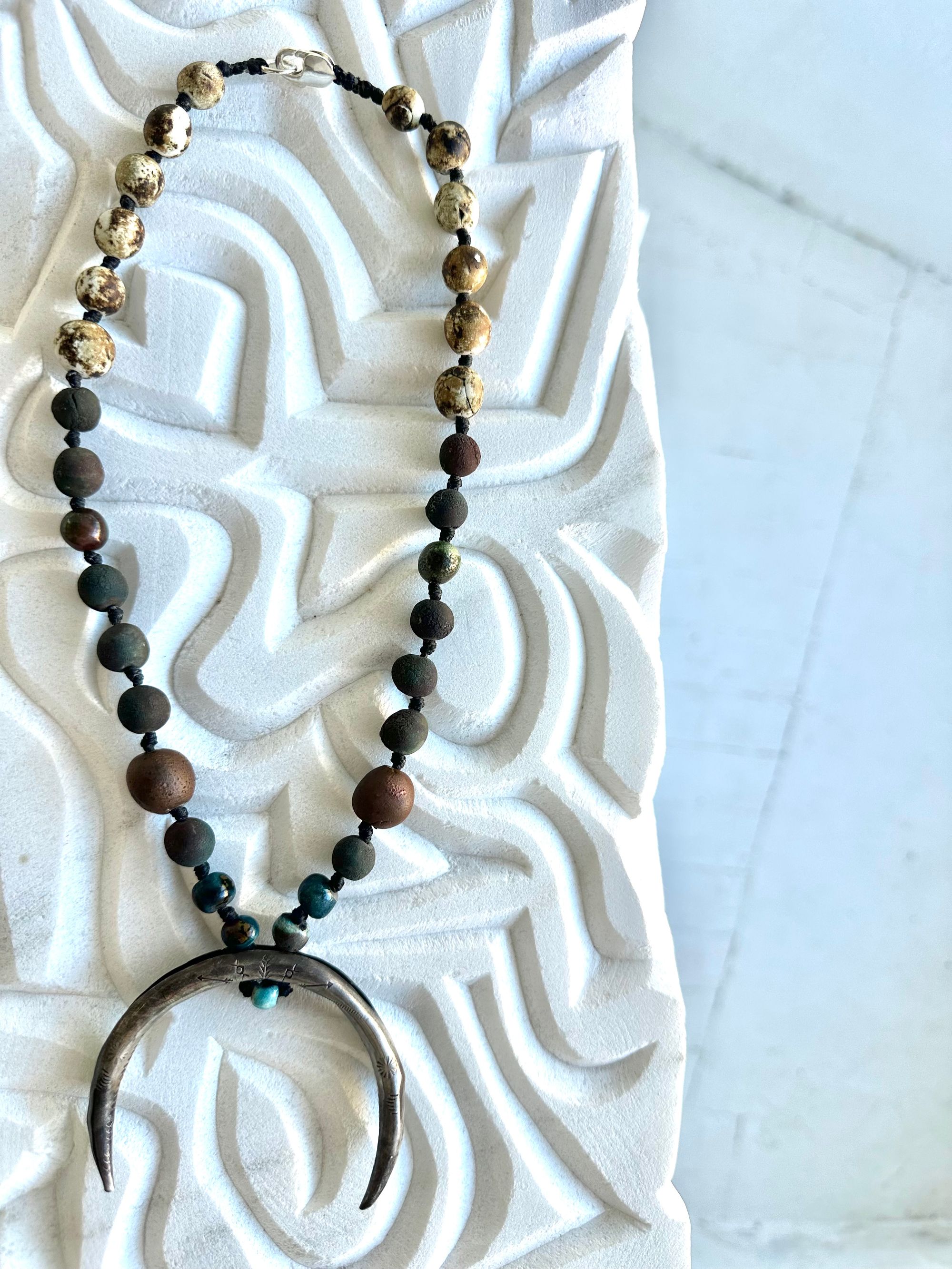 a necklace with a silver crescent moon and clay beads in earthy brown, blue and gray colors hangs from an abstract marble sculpture. The beads look like lava or stone