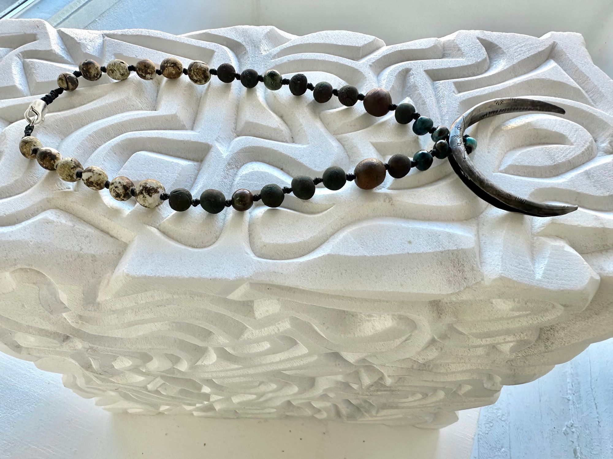 a necklace with a silver crescent moon and clay beads in earthy brown, blue and gray colors hangs from an abstract marble sculpture. The beads look like lava or stone