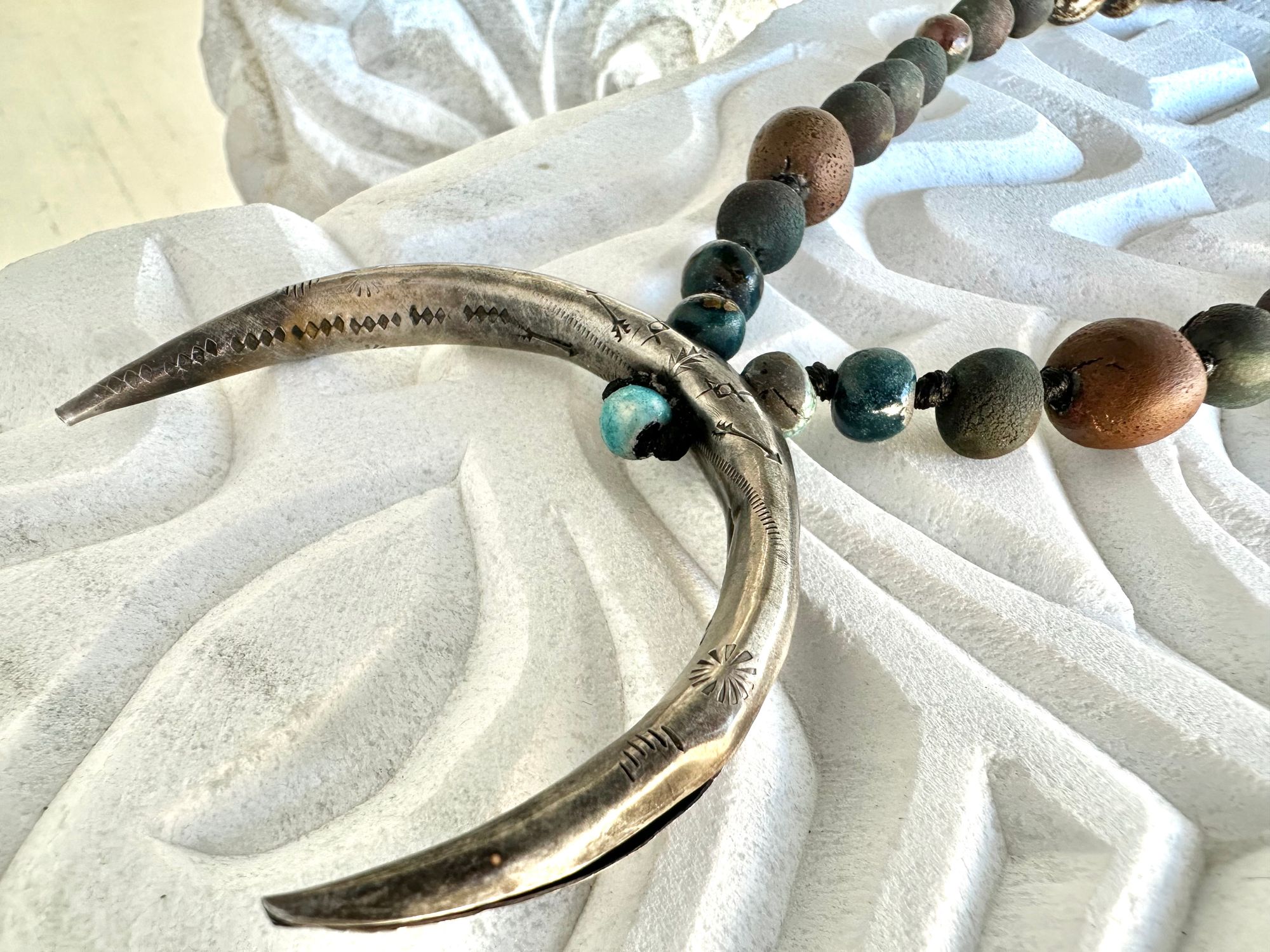 a necklace with a silver crescent moon and clay beads in earthy brown, blue and gray colors hangs from an abstract marble sculpture. The beads look like lava or stone