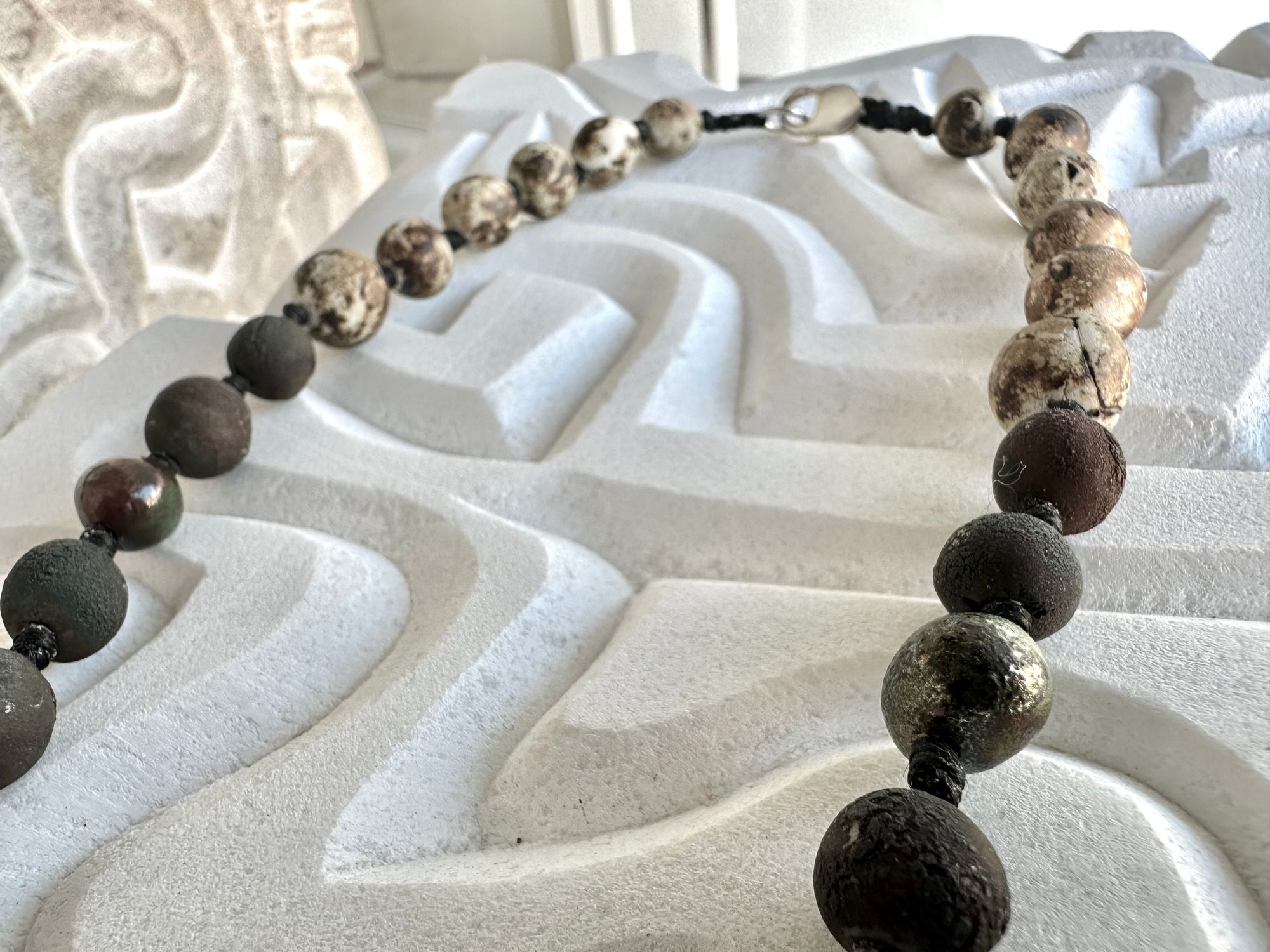 a necklace with a silver crescent moon and clay beads in earthy brown, blue and gray colors hangs from an abstract marble sculpture. The beads look like lava or stone