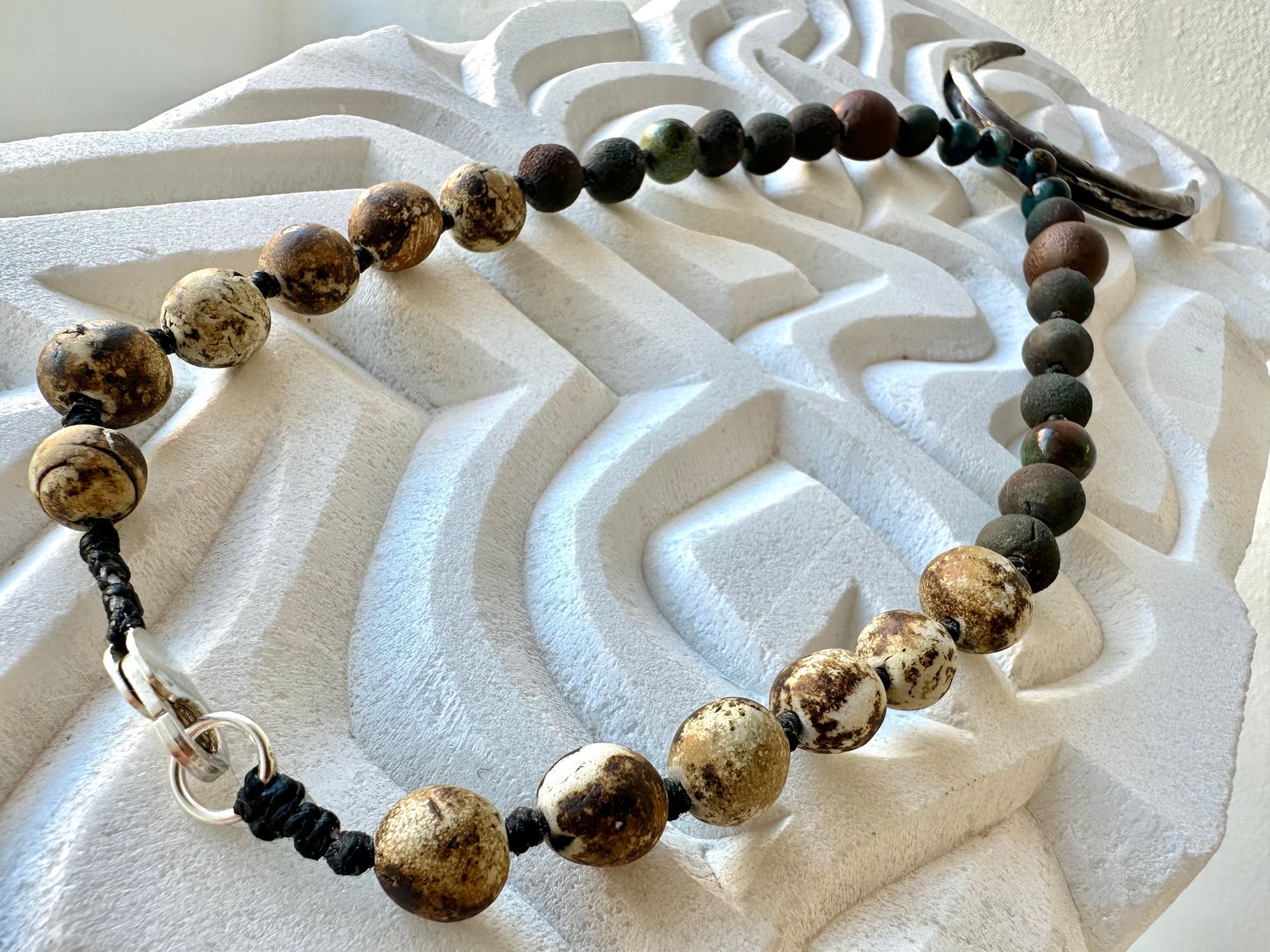 a necklace with a silver crescent moon and clay beads in earthy brown, blue and gray colors hangs from an abstract marble sculpture. The beads look like lava or stone