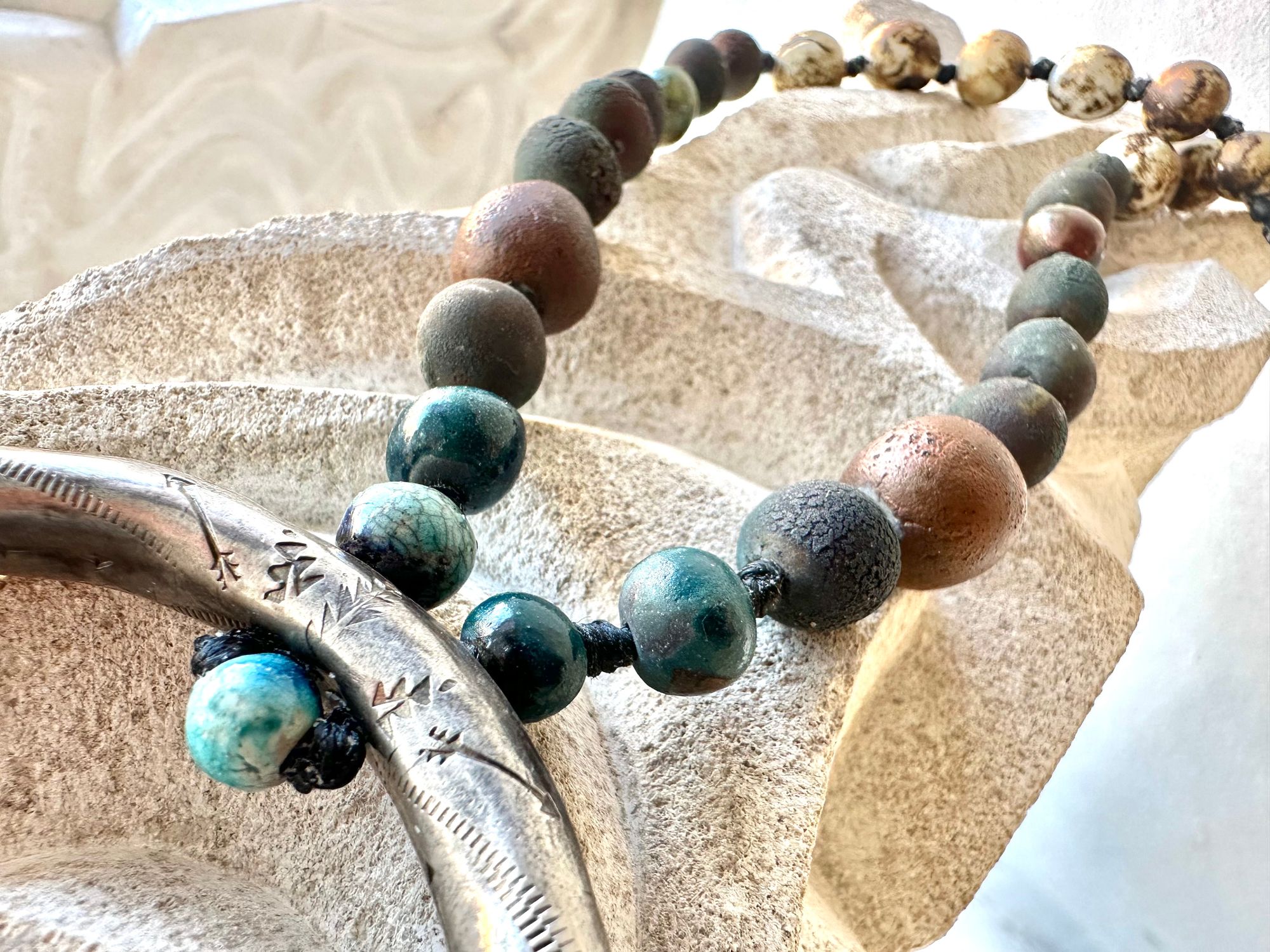 a necklace with a silver crescent moon and clay beads in earthy brown, blue and gray colors hangs from an abstract marble sculpture. The beads look like lava or stone